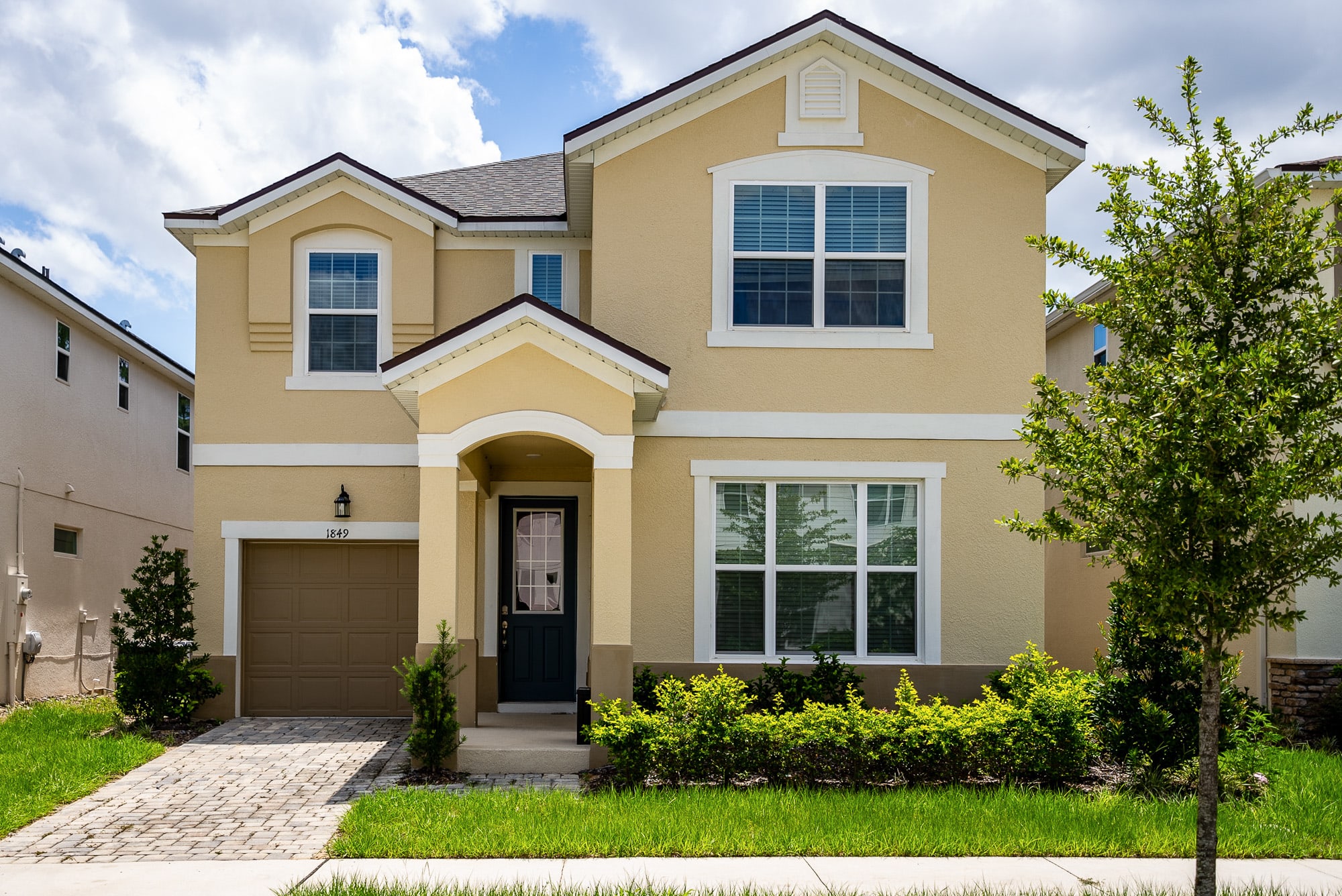 A two-story beige house with a brown roof and white trim features a covered front entrance, a single-car garage, and a paved driveway, surrounded by green grass and manicured shrubs under a blue sky.