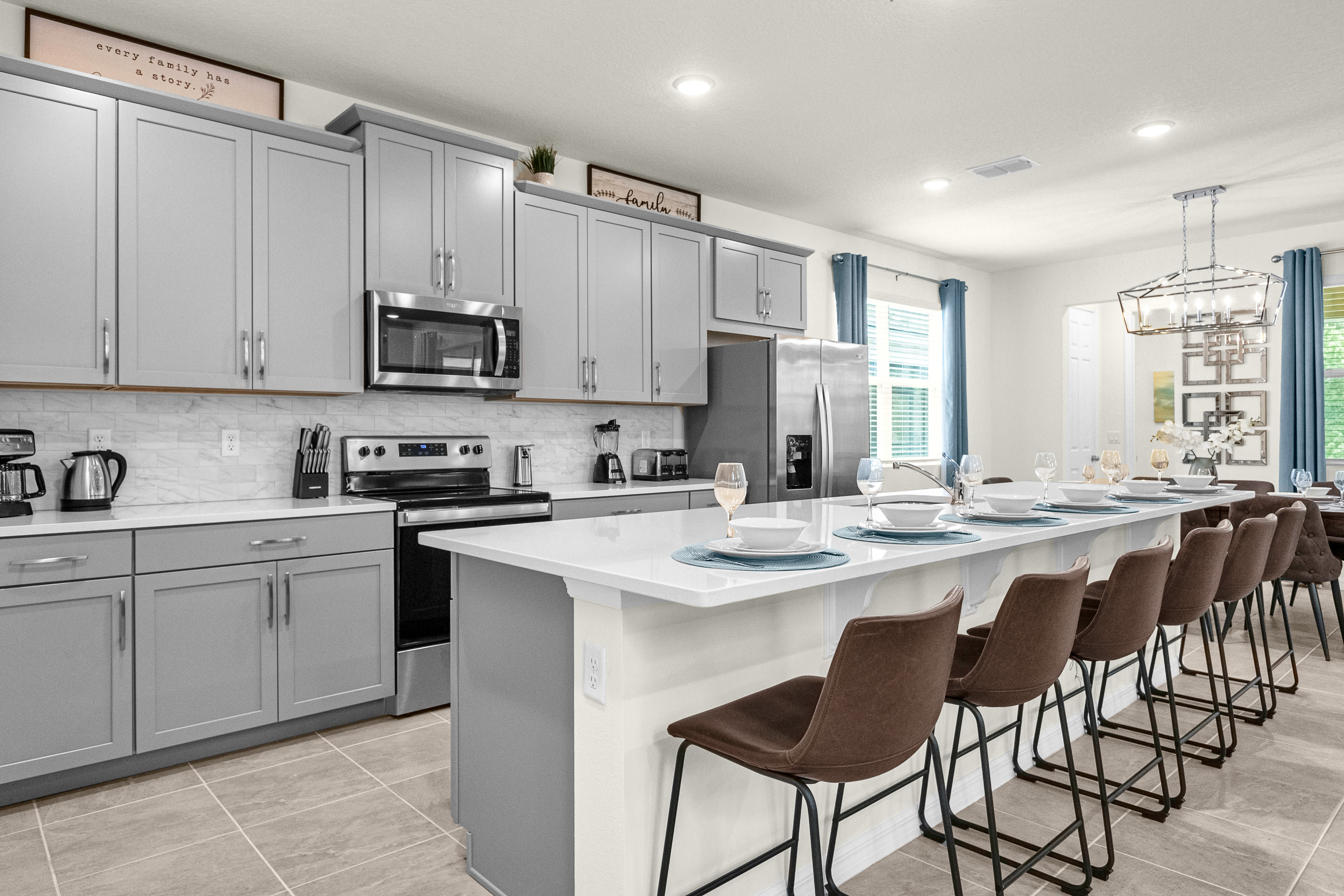 Modern kitchen with light gray shaker cabinets, white quartz countertops, stainless steel appliances, and a large island with four bar stools, connecting to the dining area.