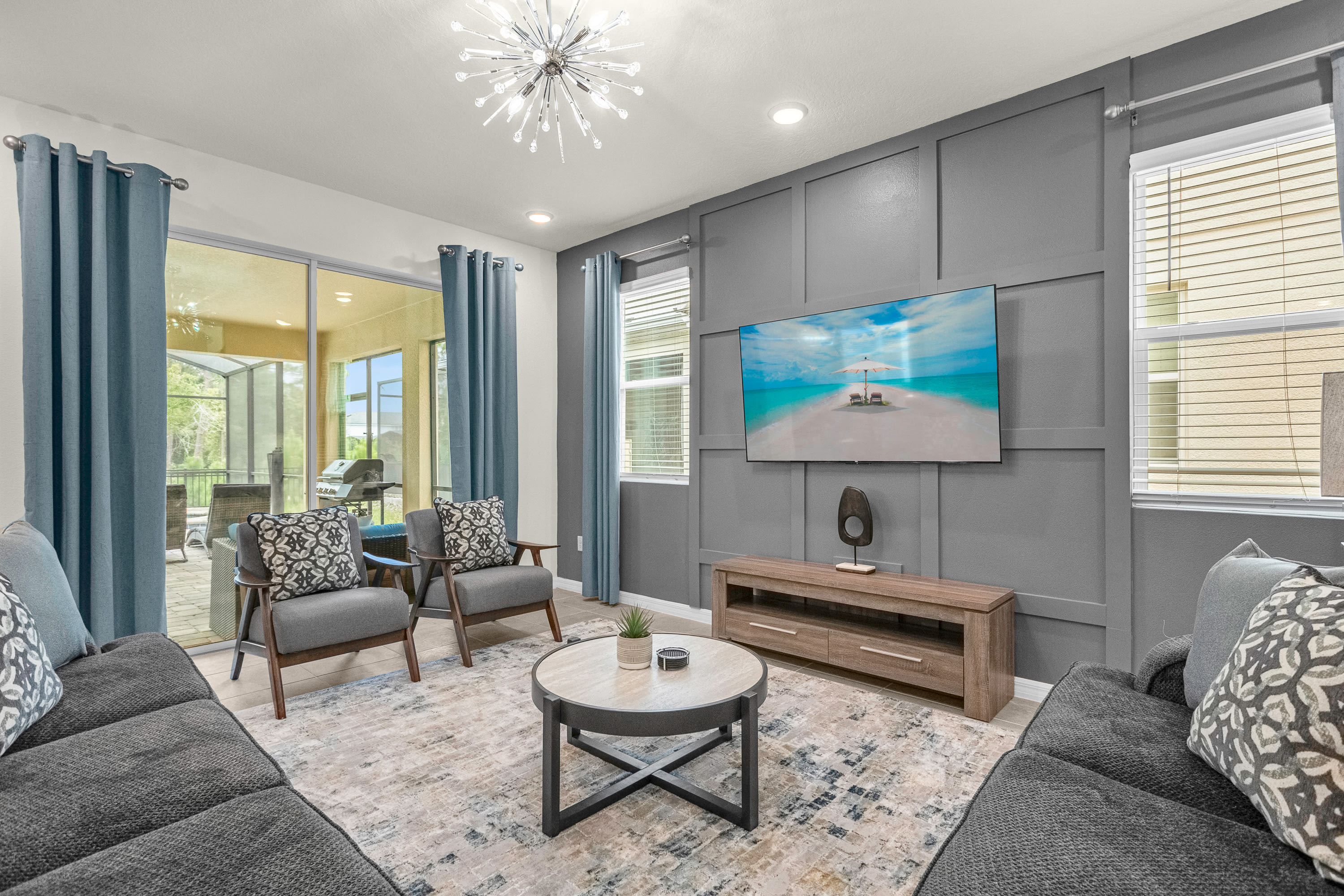 Spacious living room featuring a large gray sectional sofa, two accent chairs, a round coffee table, and a wall-mounted TV, all set on a patterned area rug. Sliding glass doors open to an outdoor patio.