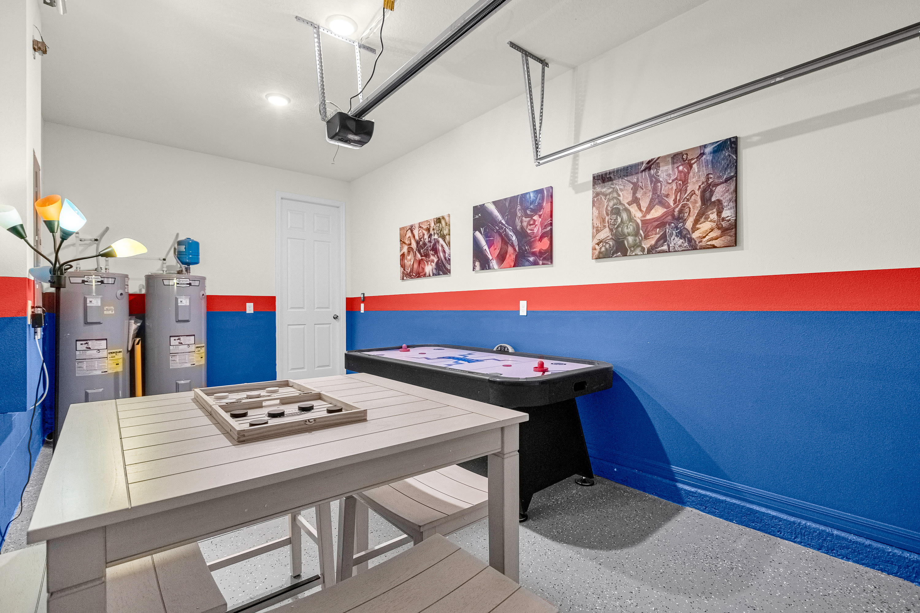A vibrant game room in a garage conversion features an air hockey table, a light wood dining table with benches, and colorful superhero artwork on walls painted with red and blue stripes.