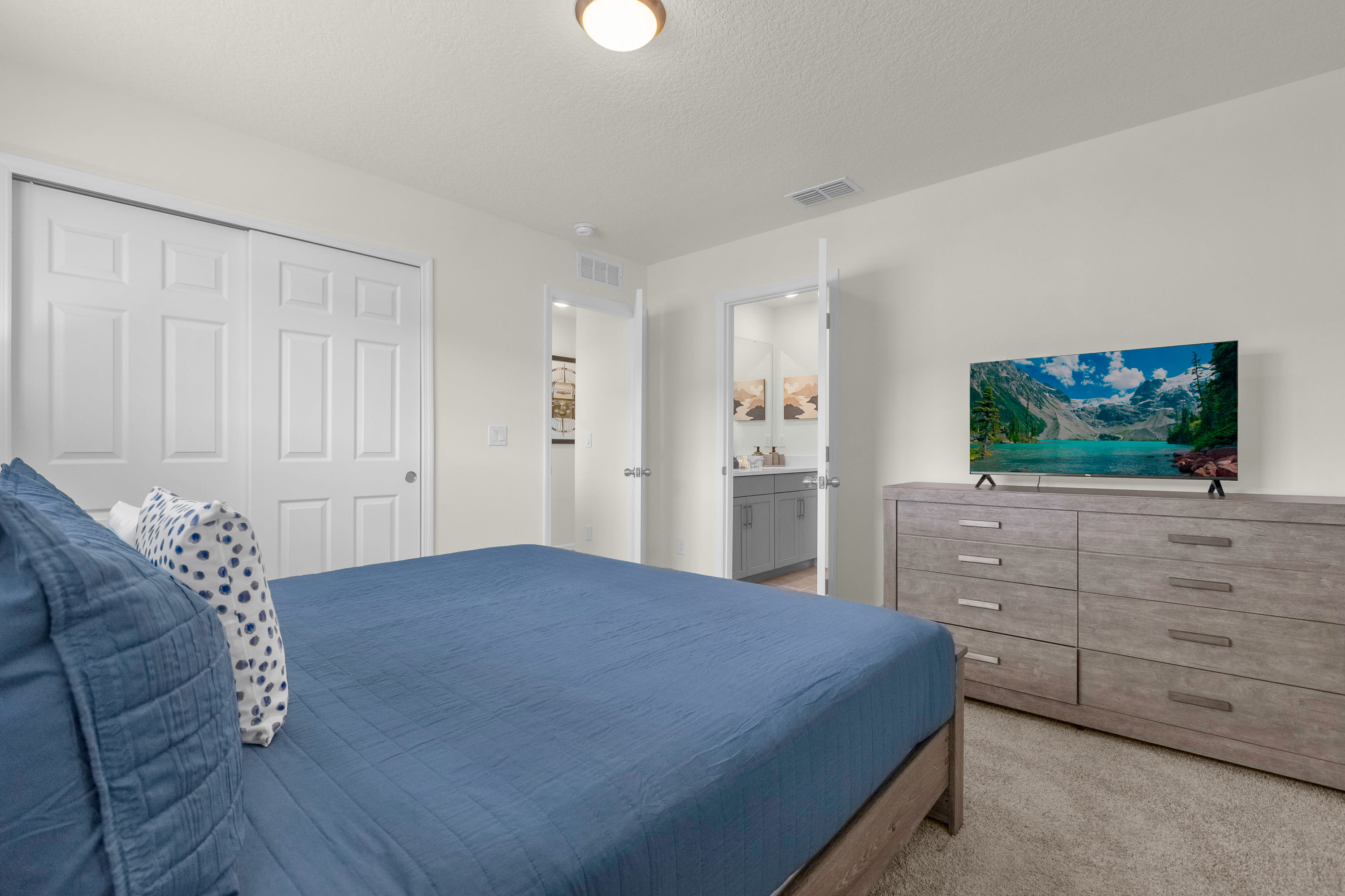 A serene bedroom featuring a large bed with a blue comforter and patterned pillows, a light wood dresser with a television, and a glimpse into an en-suite bathroom with a vanity.