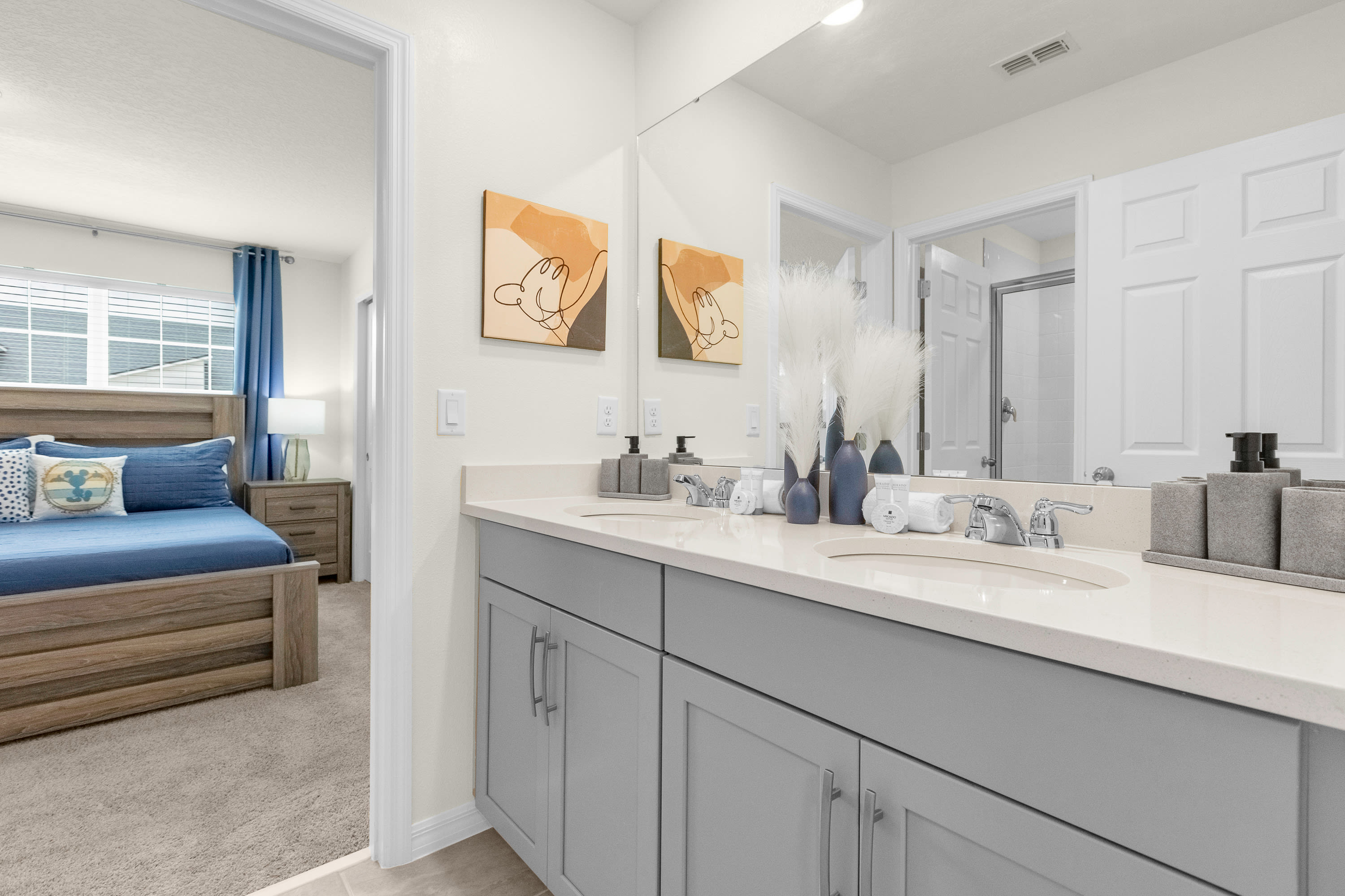 A modern en-suite bathroom with a double vanity featuring light gray cabinets, white countertops, and two sinks, adorned with abstract art and decorative pampas grass. A bedroom is visible through the doorway.