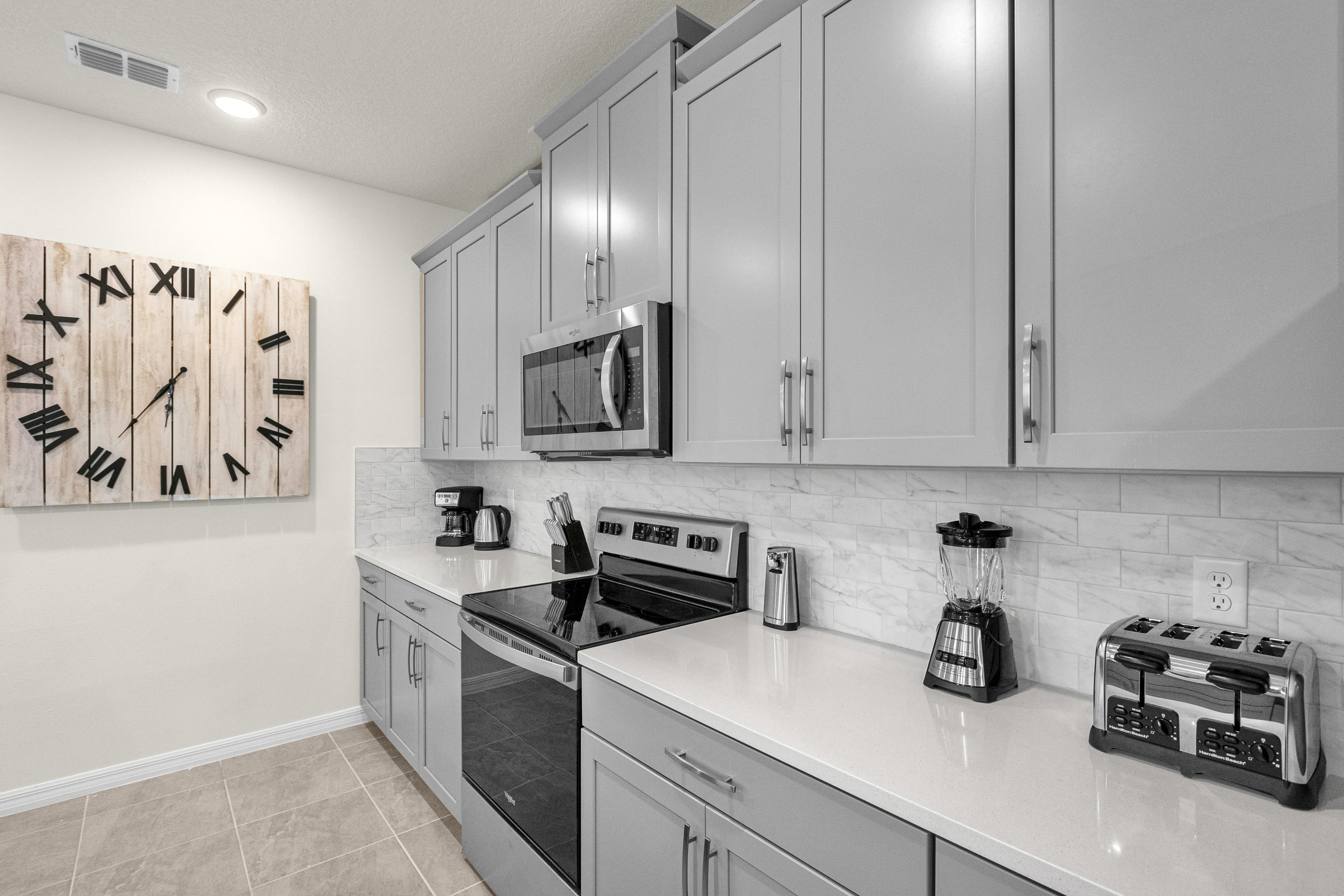 Detailed view of the kitchen counter showcasing stainless steel appliances including a microwave, stove, toaster, blender, and coffee maker, with a white subway tile backsplash and a large wooden clock on the wall.