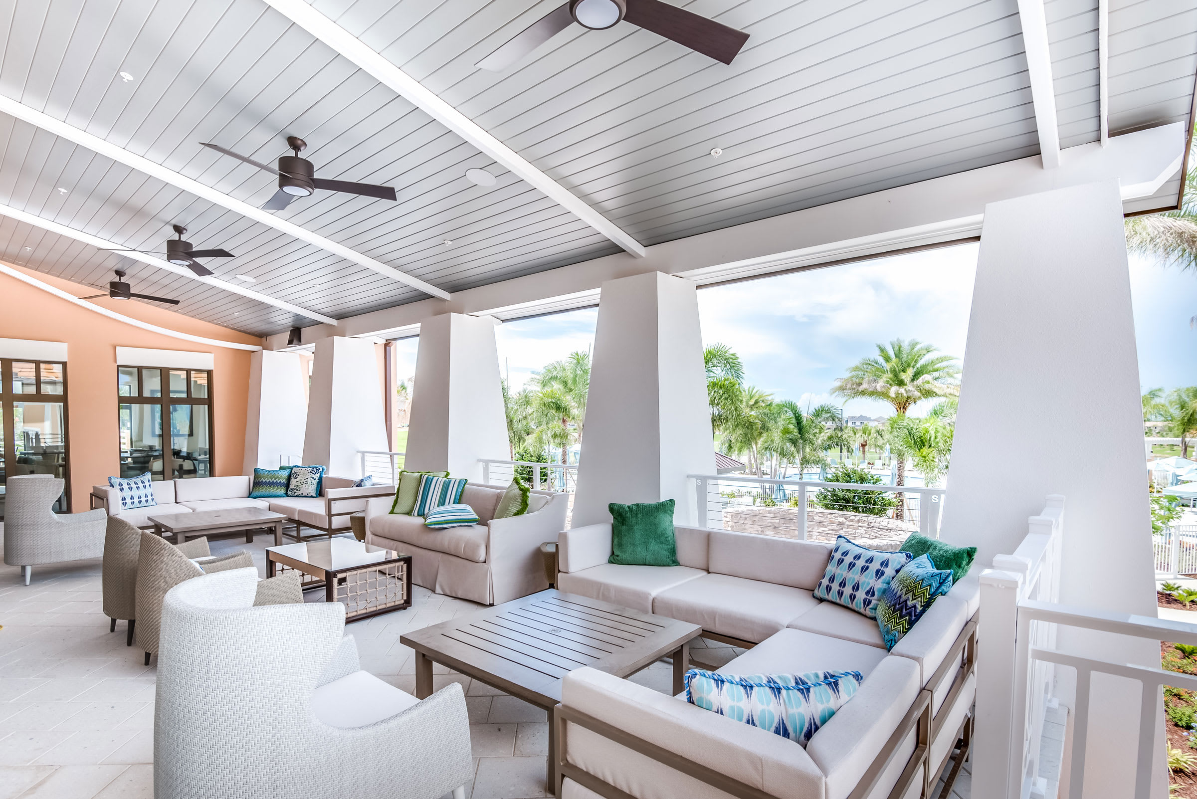 A stylish covered outdoor lounge area featuring comfortable sofas and armchairs with decorative pillows, coffee tables, and ceiling fans, offering views of a pool area and palm trees.