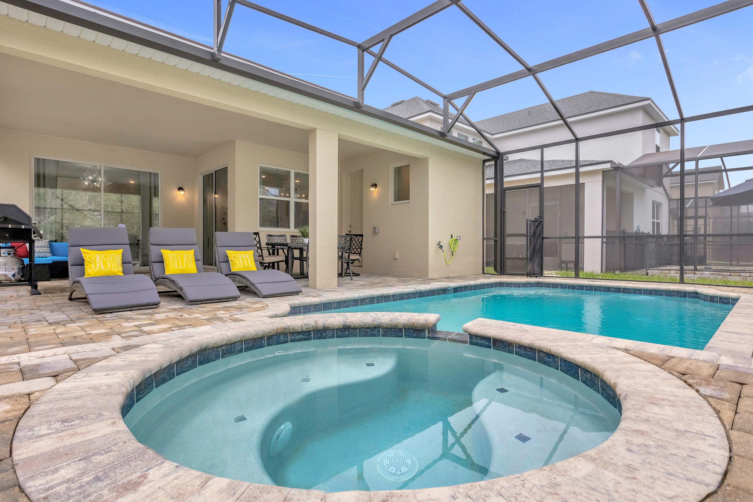 A screened-in outdoor pool area features a large hot tub in the foreground and a rectangular swimming pool, surrounded by a paved deck with three grey lounge chairs and a covered patio with a dining set.