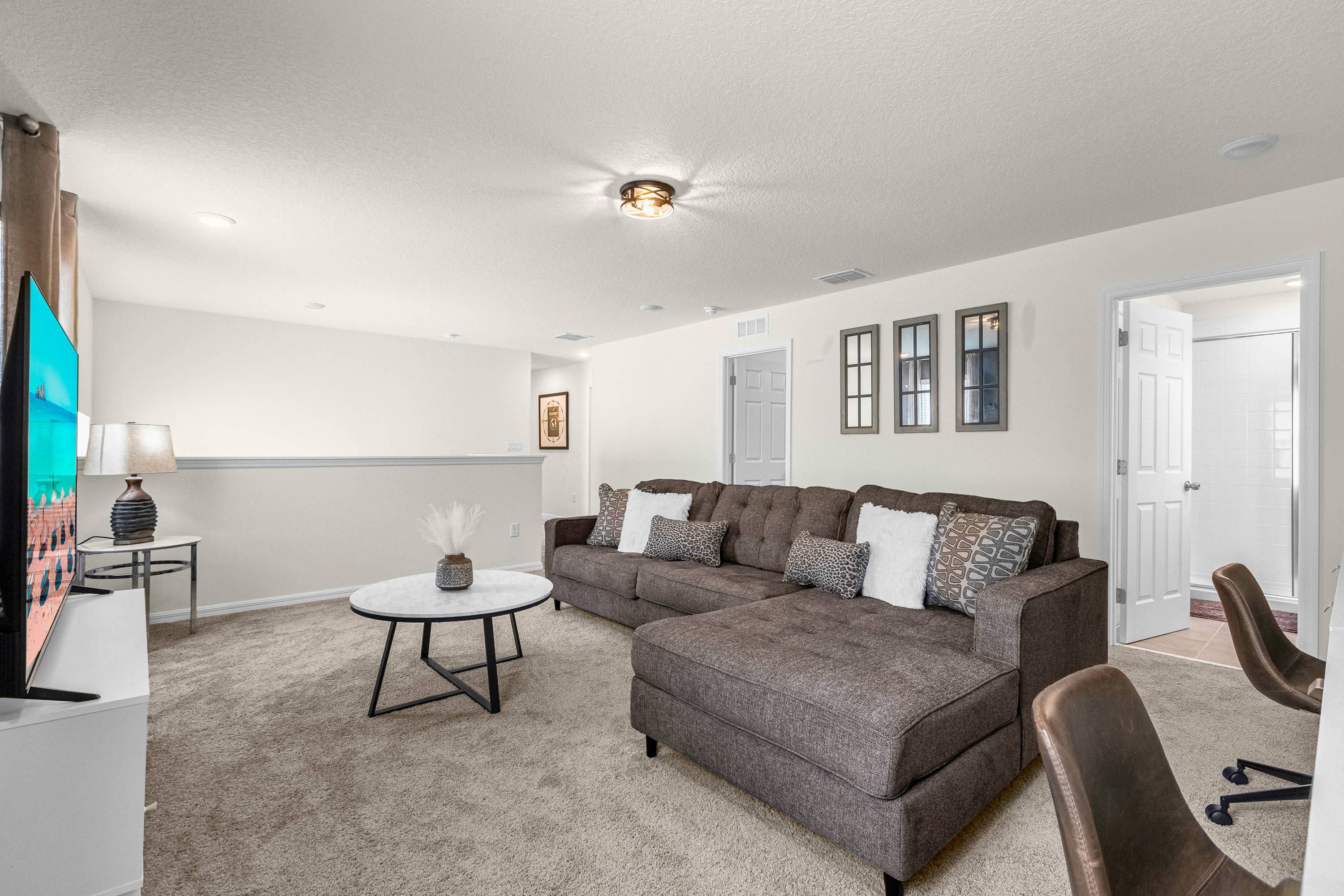 Upstairs secondary living area featuring a large brown sectional sofa, a round coffee table, a wall-mounted TV, and carpeted flooring, with a dedicated workspace visible in the background.