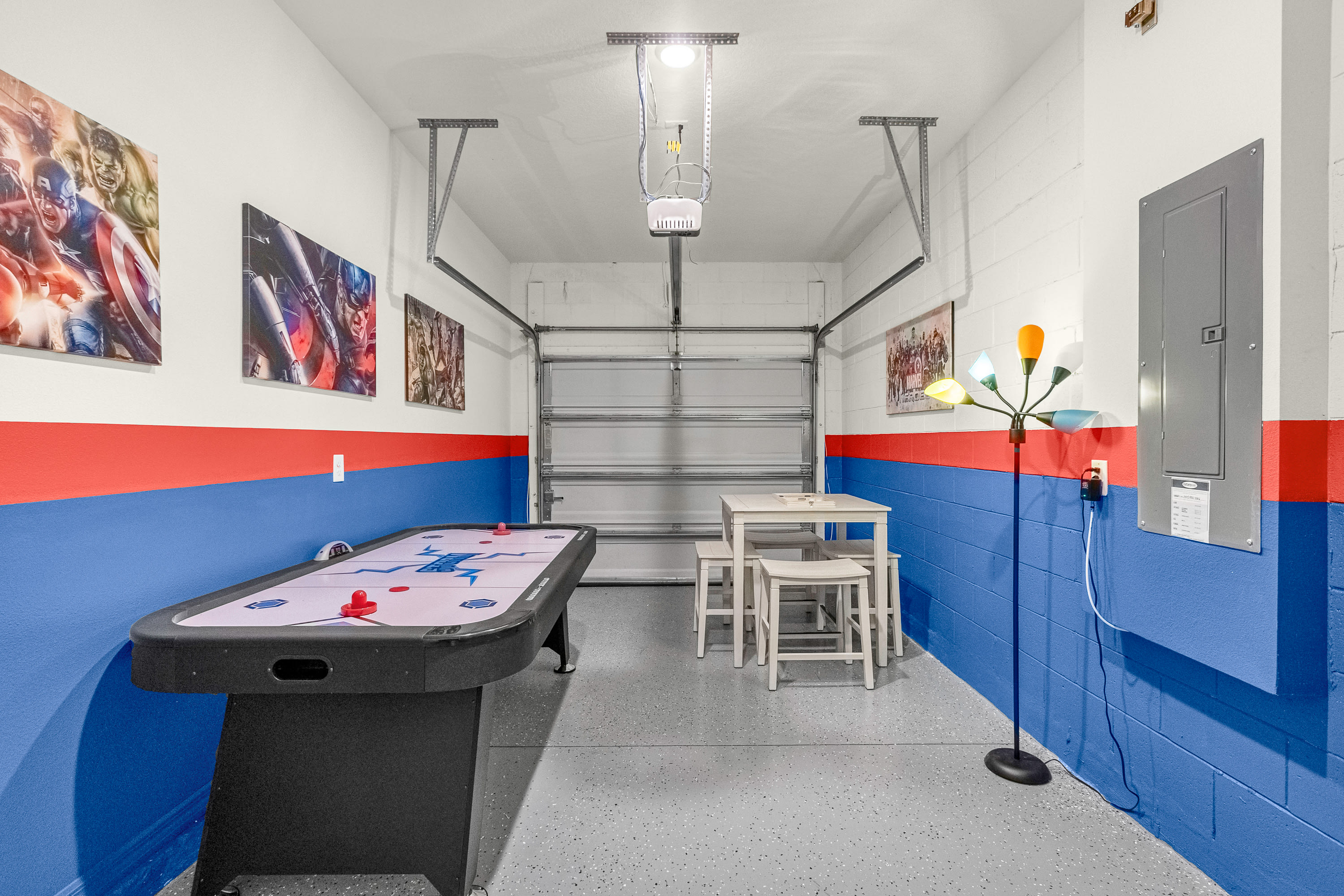 A garage converted into a game room features an air hockey table, a small white table with stools, and colorful superhero artwork on walls painted with red and blue stripes, with the garage door visible.