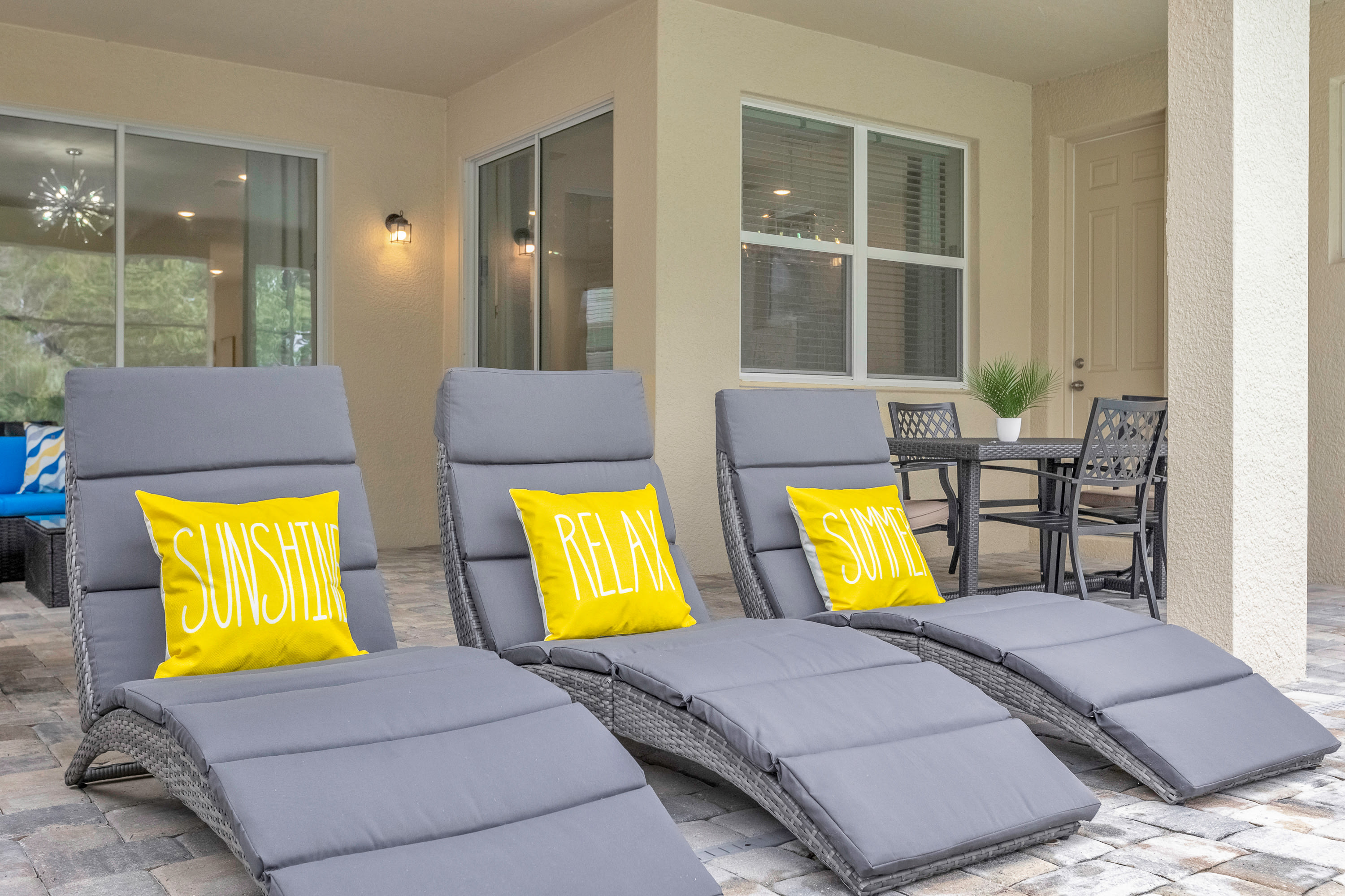 A close-up of three grey outdoor lounge chairs with bright yellow "SUNSHINE", "RELAX", and "SUMMER" pillows, positioned on a paved patio under a covered area, with a glimpse of an outdoor dining set.