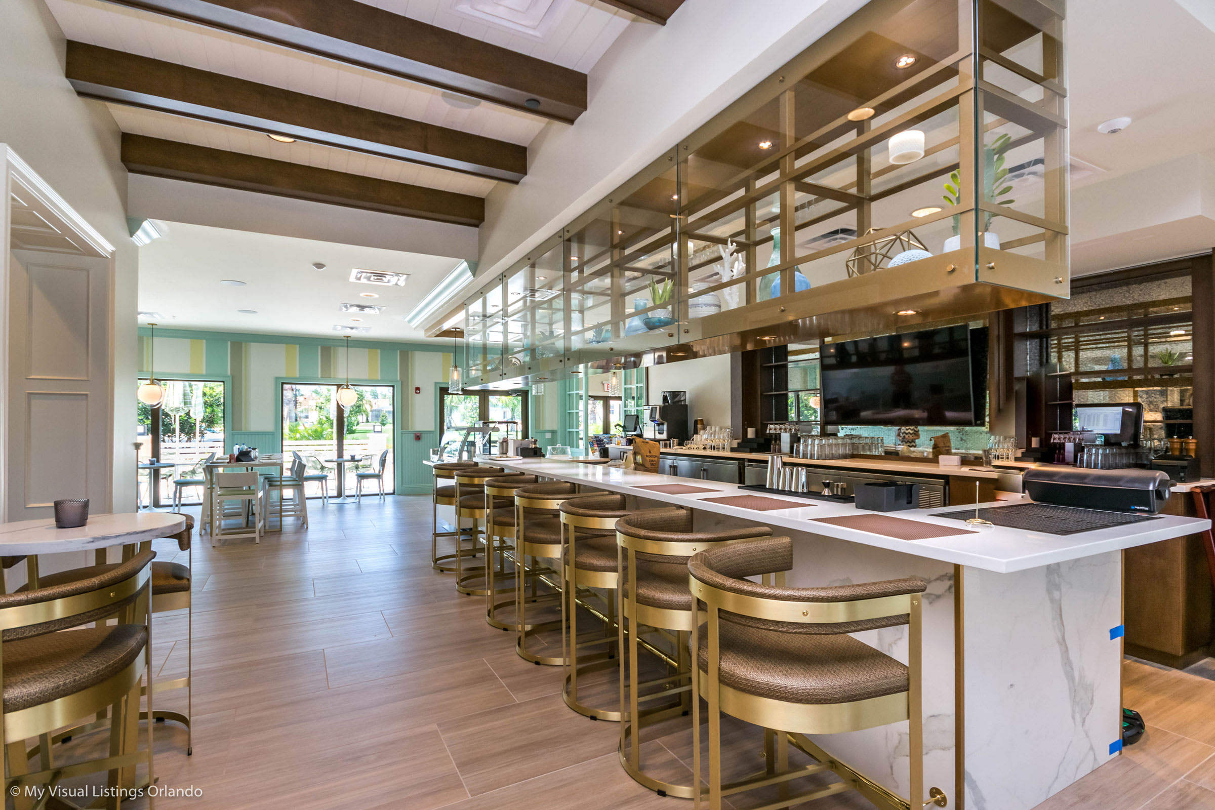 A sophisticated bar area with a long white marble counter, gold-accented bar stools, and an overhead shelving unit, leading into a bright dining space with light wood floors.