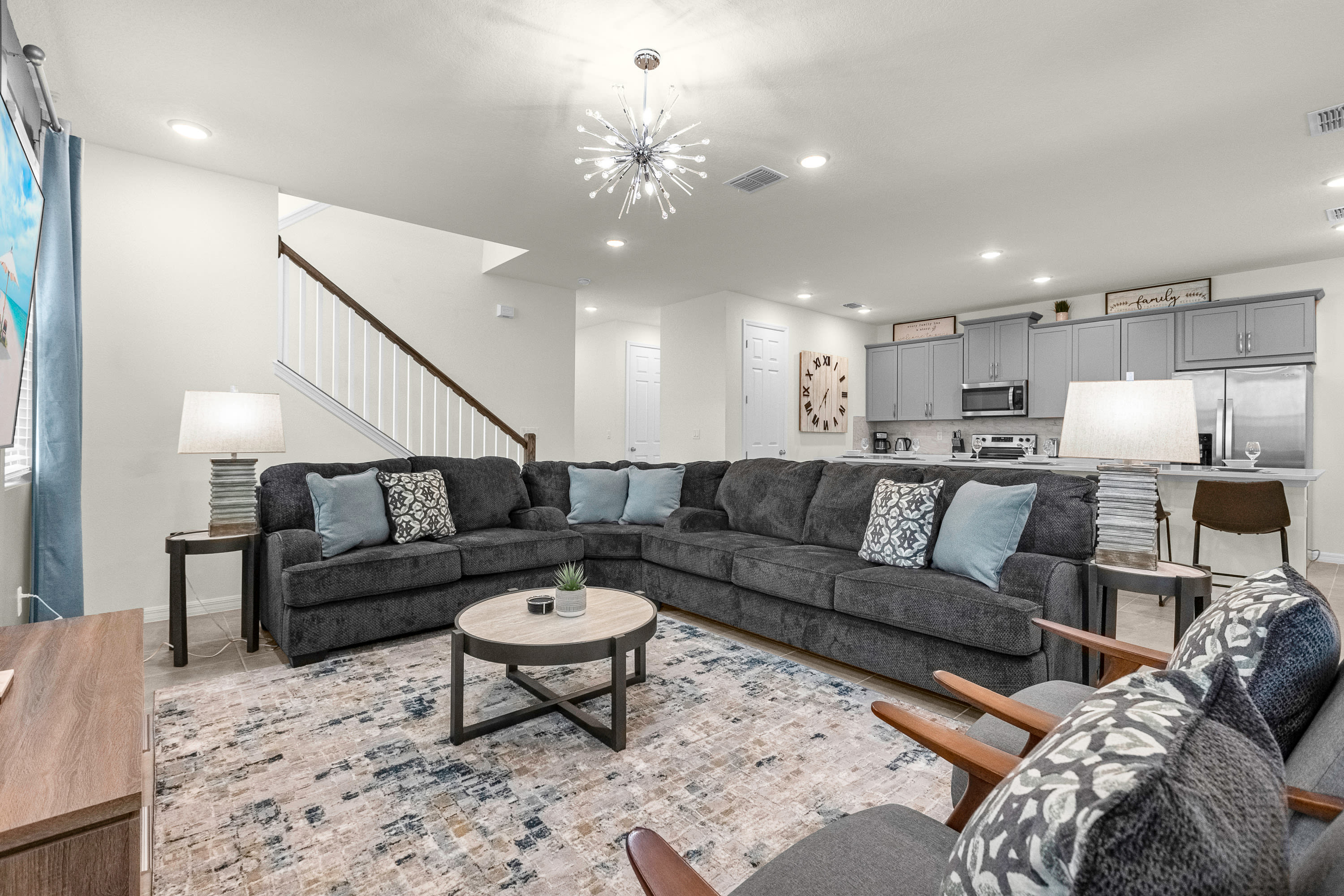 An open-concept living room features a large dark grey sectional sofa with light blue throw pillows, a round coffee table, and a patterned area rug, flowing into a modern grey kitchen with a breakfast bar.