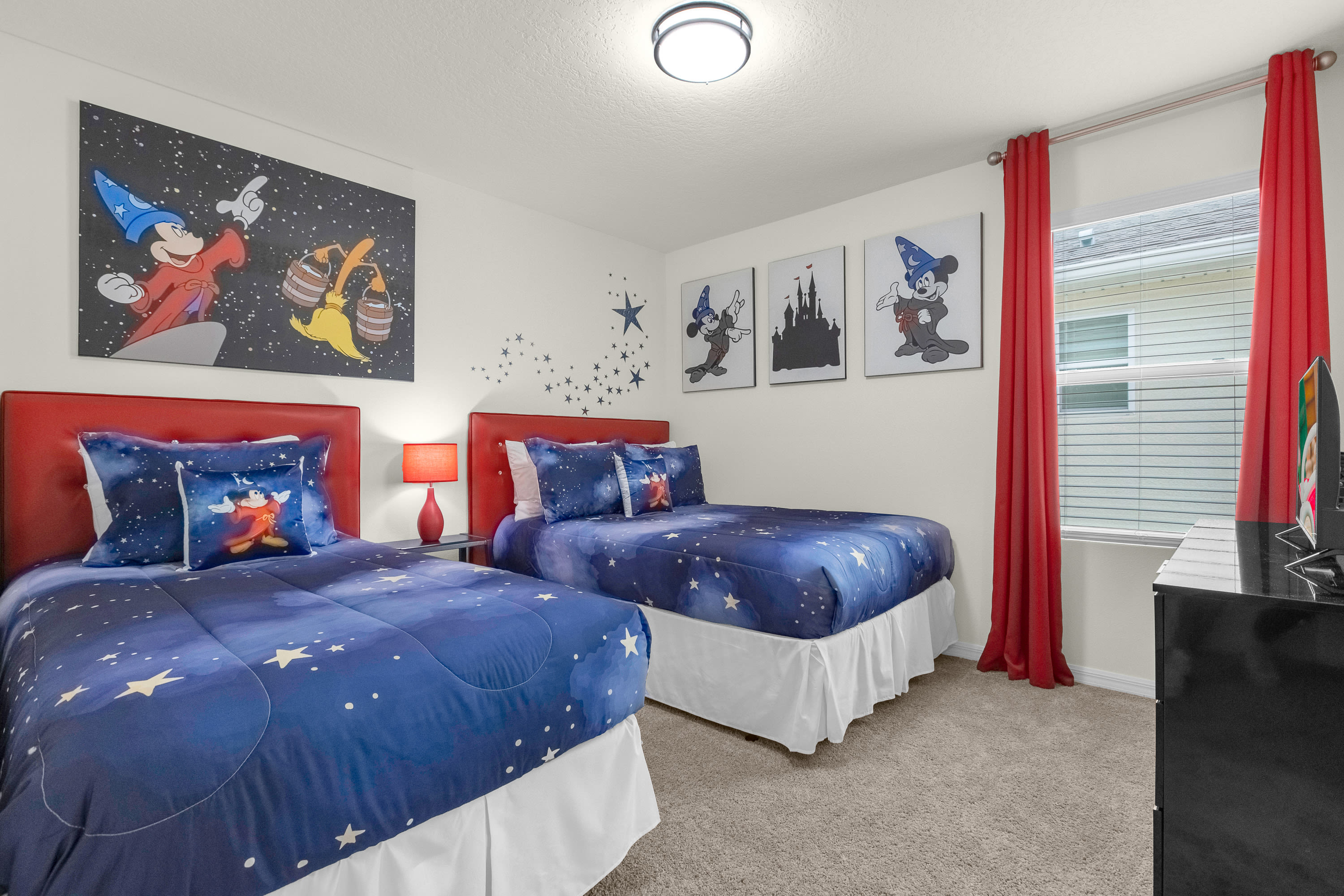 A vibrant, Disney-themed bedroom with two twin beds featuring red headboards and blue star-patterned bedding, adorned with large Mickey Mouse artwork and red curtains.