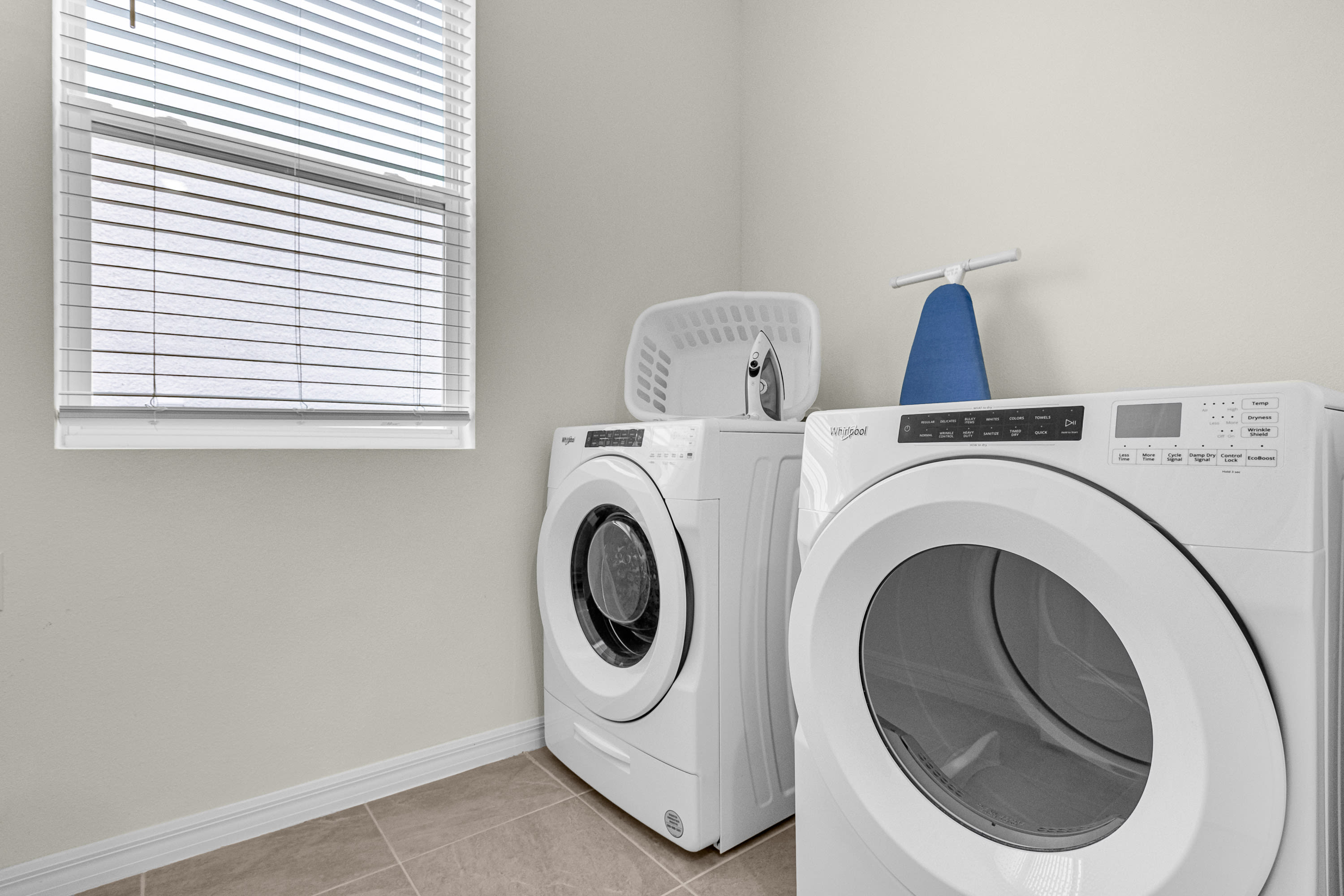 Functional laundry room equipped with a white front-loading Whirlpool washer and dryer set. A window with white blinds provides natural light, and a laundry basket and ironing board are conveniently placed on top of the machines.