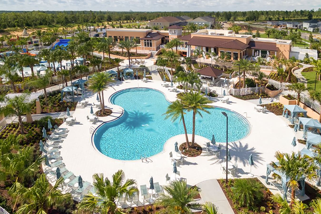 Aerial view of a sprawling resort-style communal pool complex featuring multiple interconnected pools, surrounded by numerous lounge chairs, umbrellas, and lush palm trees. Several resort buildings are visible in the background.