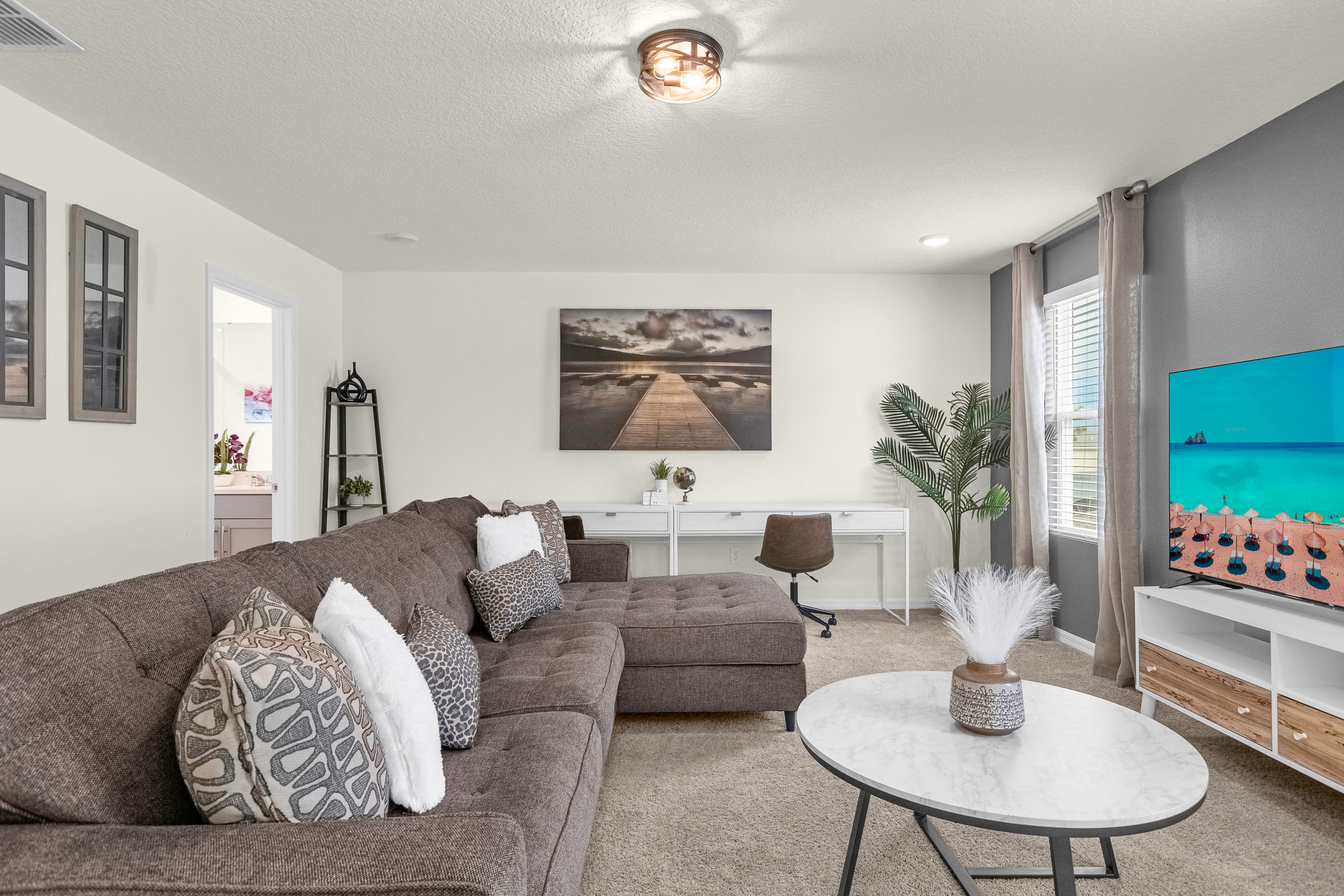 A bright upstairs living area features a large brown sectional sofa with decorative pillows, a white marble-top coffee table, a white desk with a chair, and a wall-mounted TV, all on a beige carpet.