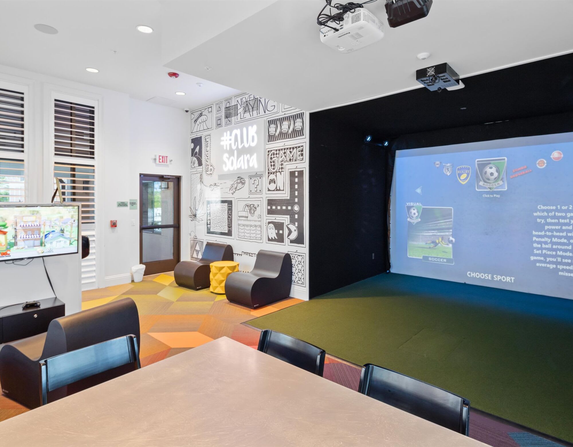 Spacious indoor game room or clubhouse lounge area featuring a golf simulator screen, a large TV, and various seating options including modern armchairs and a dining table. A graphic mural decorates one wall.