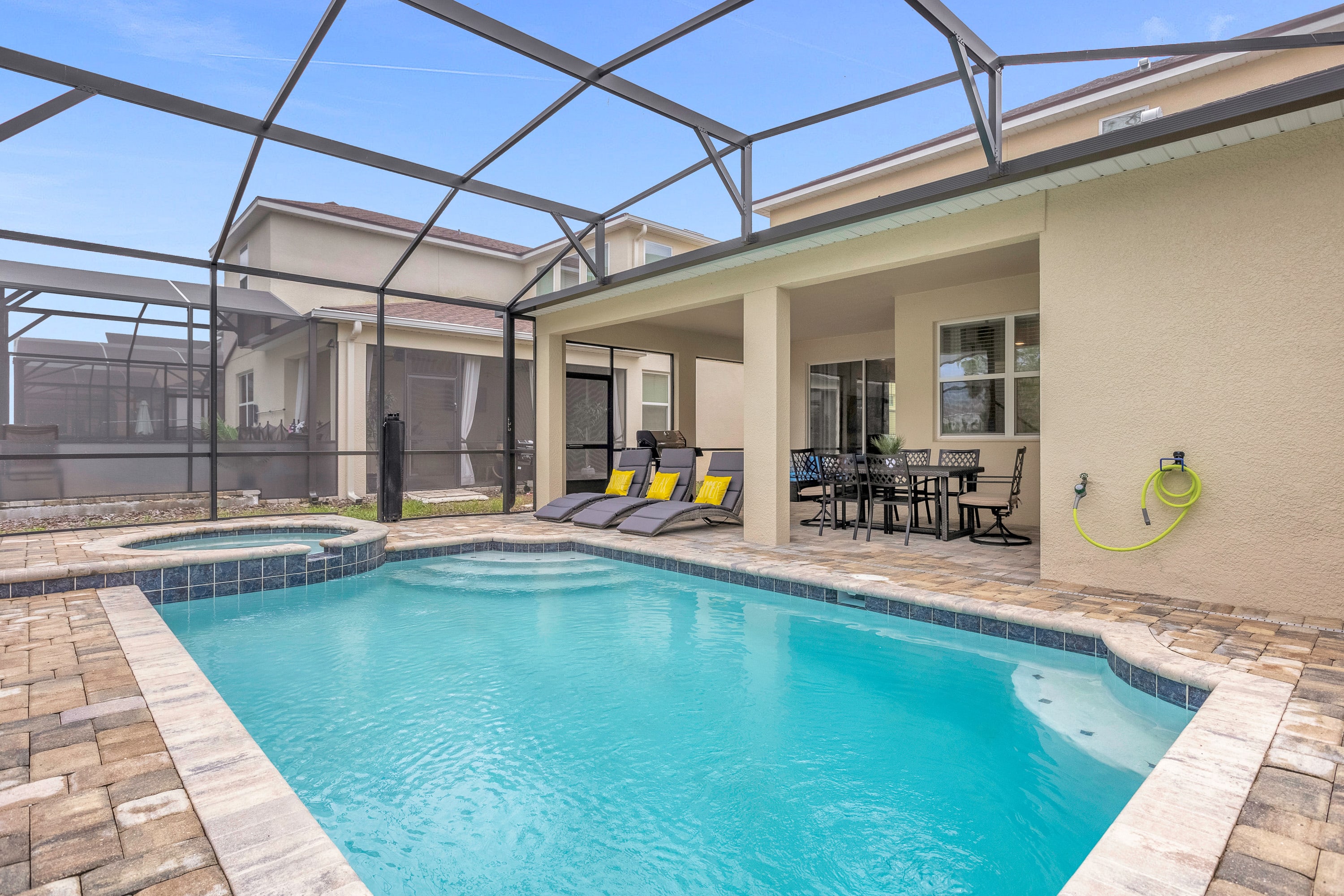 A screened-in outdoor pool area features a rectangular swimming pool with steps, an integrated hot tub, and two grey lounge chairs with yellow pillows on a paved deck, all under a clear blue sky.