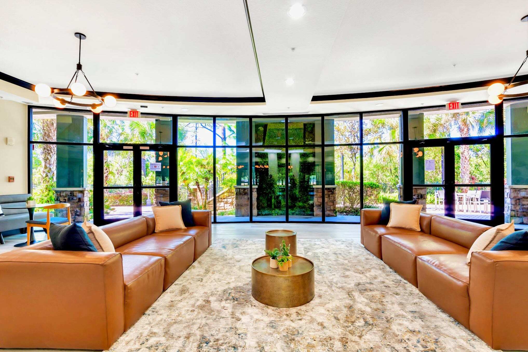 A modern and spacious communal lobby area featuring two large brown leather sofas, two round metallic gold coffee tables, and a light-colored patterned rug, all set against large floor-to-ceiling windows overlooking lush outdoor greenery.