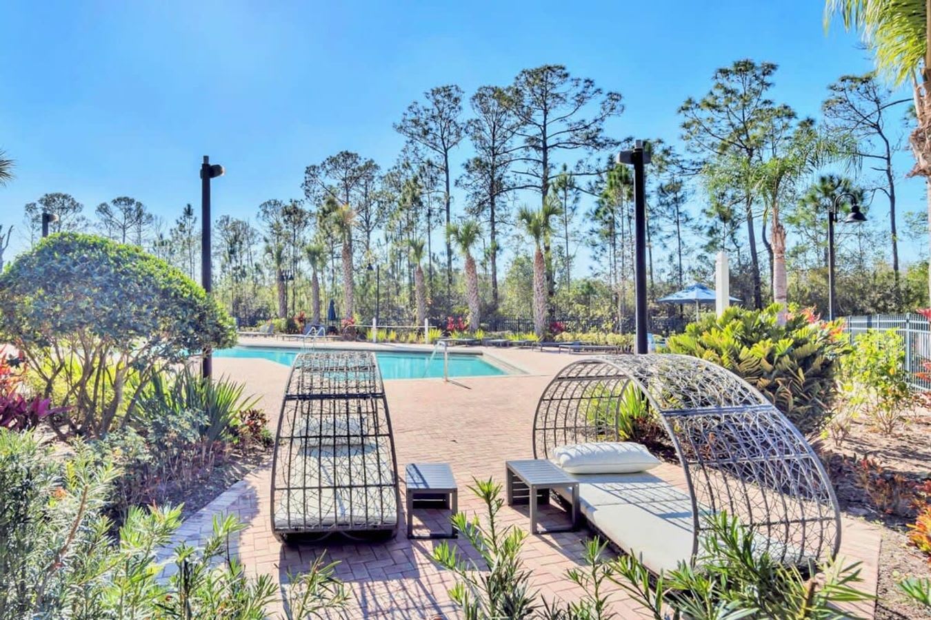 Outdoor communal pool area surrounded by lush green landscaping and tall trees under a bright blue sky. Two modern, egg-shaped lounge chairs with cushions are visible on the brick patio next to the sparkling blue pool.