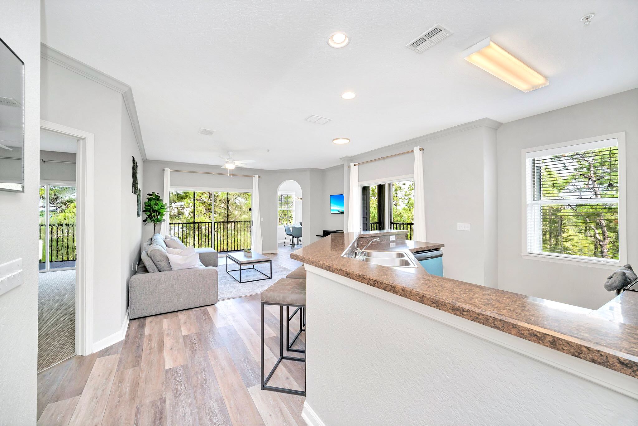 Open-concept living space featuring a light gray sofa, a modern coffee table, and a view of a balcony with lush greenery, adjacent to a kitchen bar with seating.