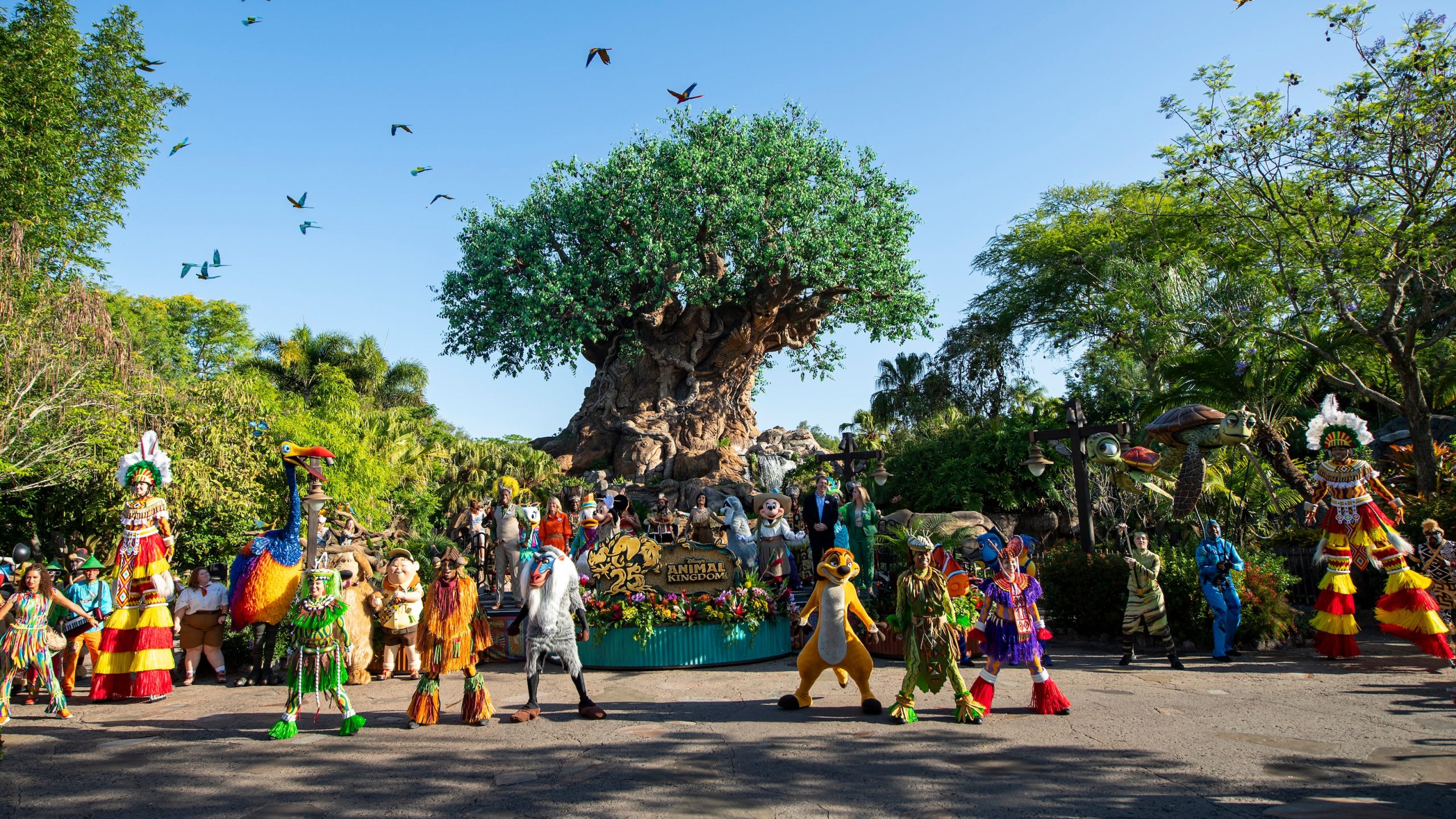 Vibrant scene at Disney's Animal Kingdom featuring the iconic Tree of Life in the background, surrounded by various costumed characters and performers. Birds fly overhead in a clear blue sky.