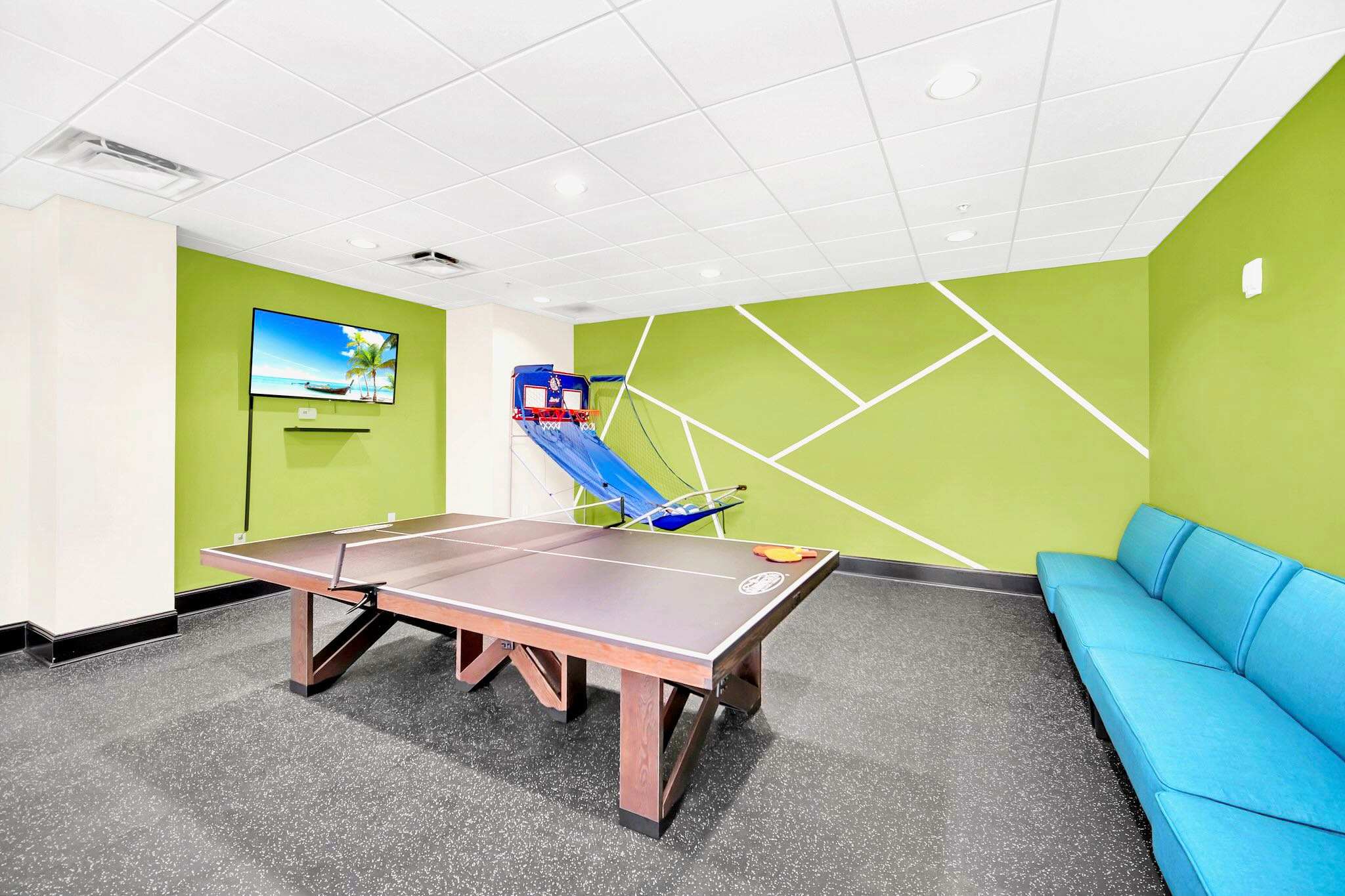 A vibrant game room equipped with a wooden ping-pong table, a basketball arcade game, a wall-mounted television, and a long blue sofa, all set against a bright green accent wall with geometric white lines.