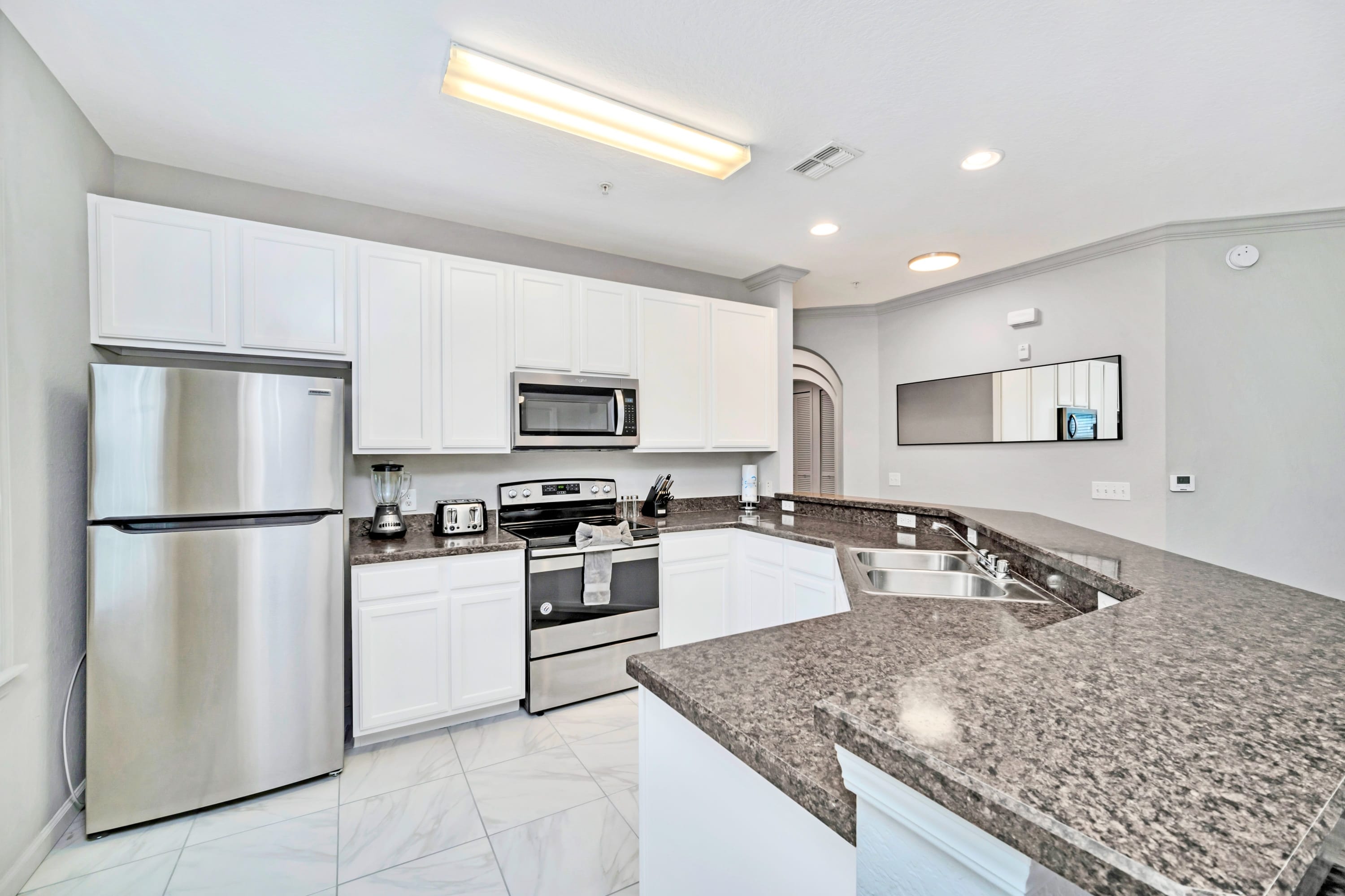 Bright kitchen featuring white shaker cabinets, stainless steel appliances including a refrigerator, microwave, and range, with dark granite countertops and white tiled flooring.