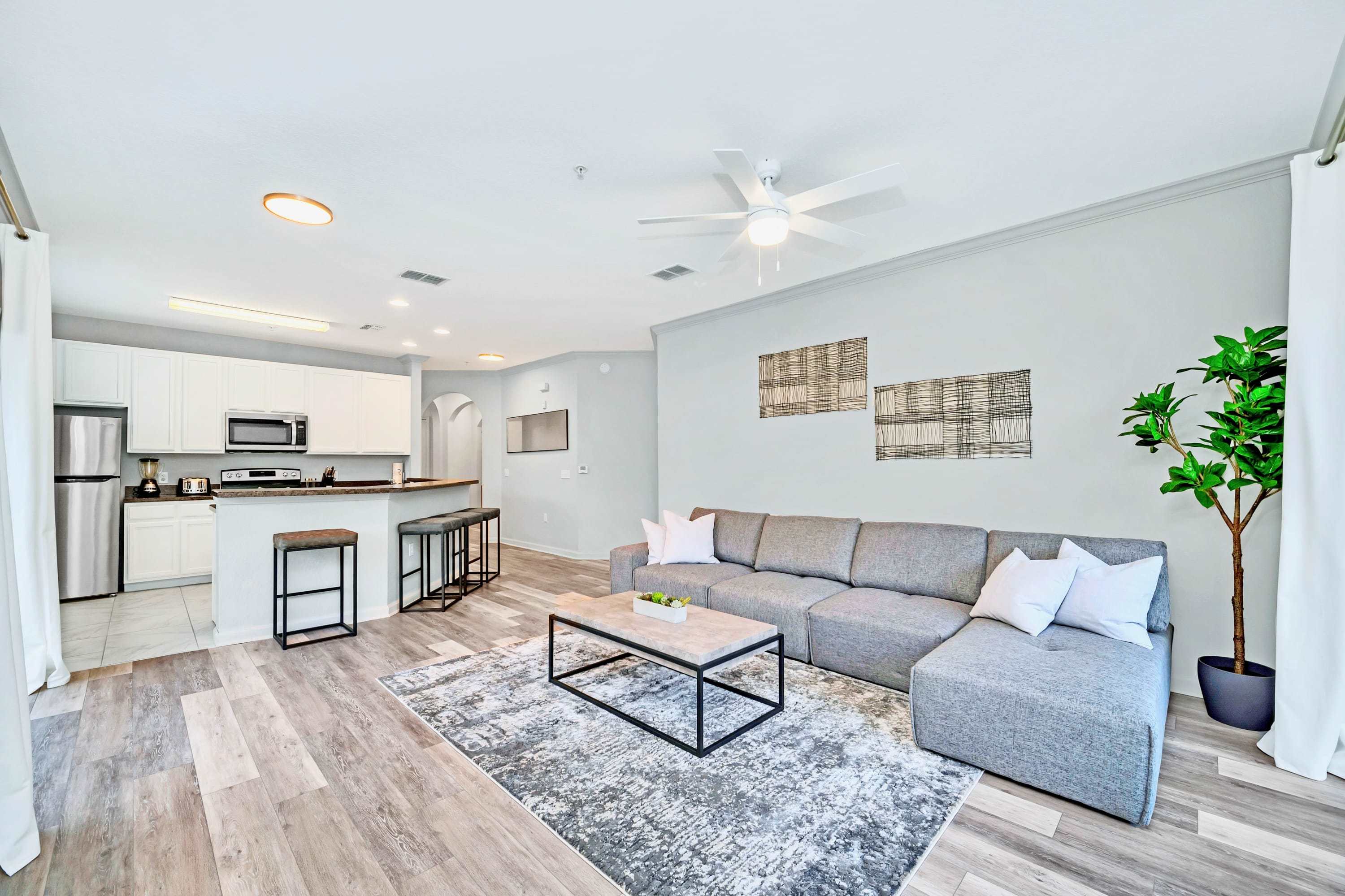Open-concept living area showing the gray L-shaped sofa, coffee table, and area rug, with a view towards the kitchen and dining area. The kitchen features white cabinetry, stainless steel appliances, and a breakfast bar with stools.