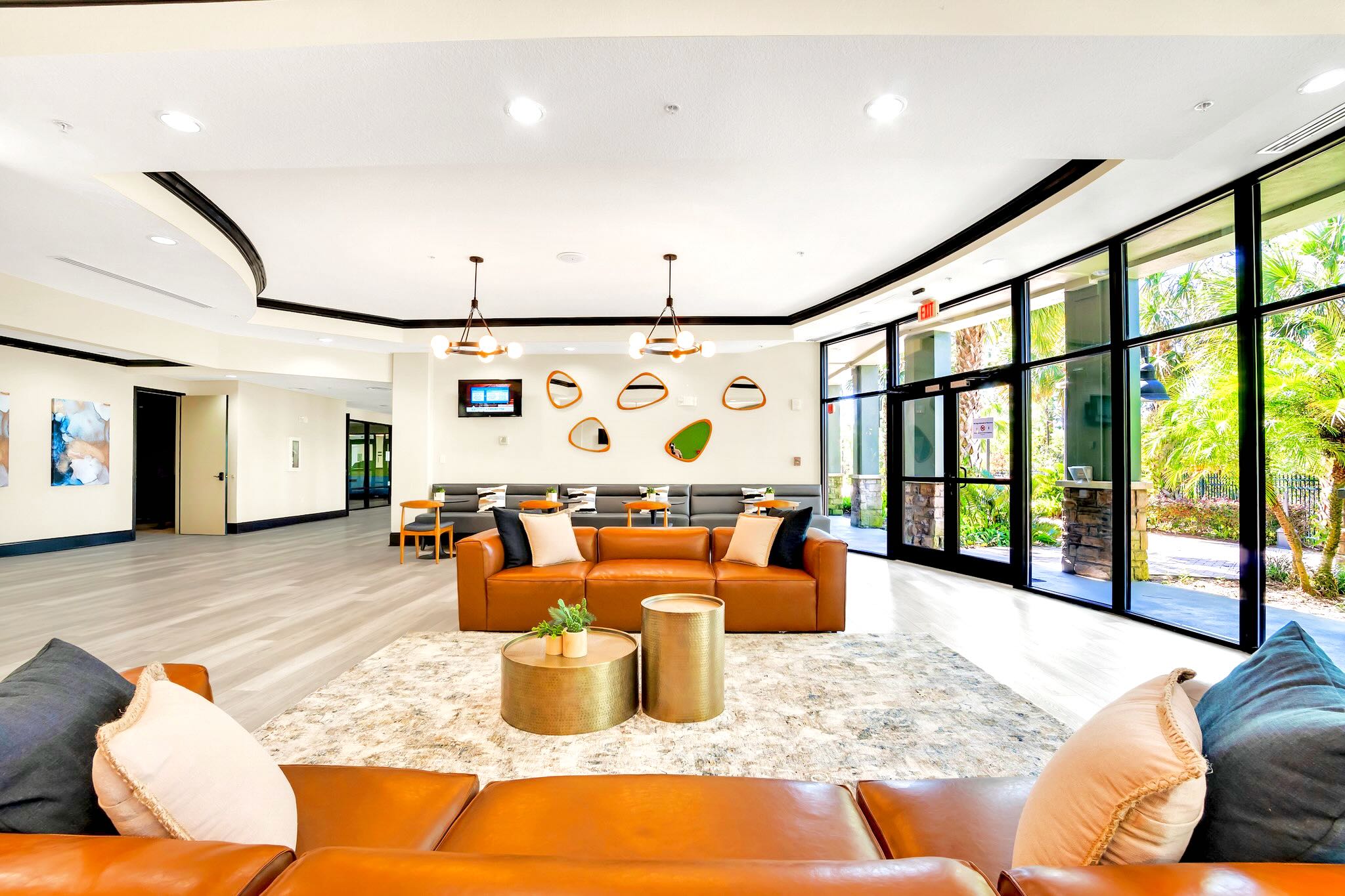 A stylish communal lounge area within a lobby, showcasing two brown leather sofas, metallic gold coffee tables, and a patterned area rug, complemented by a wall adorned with an eclectic arrangement of decorative mirrors and modern lighting fixtures.