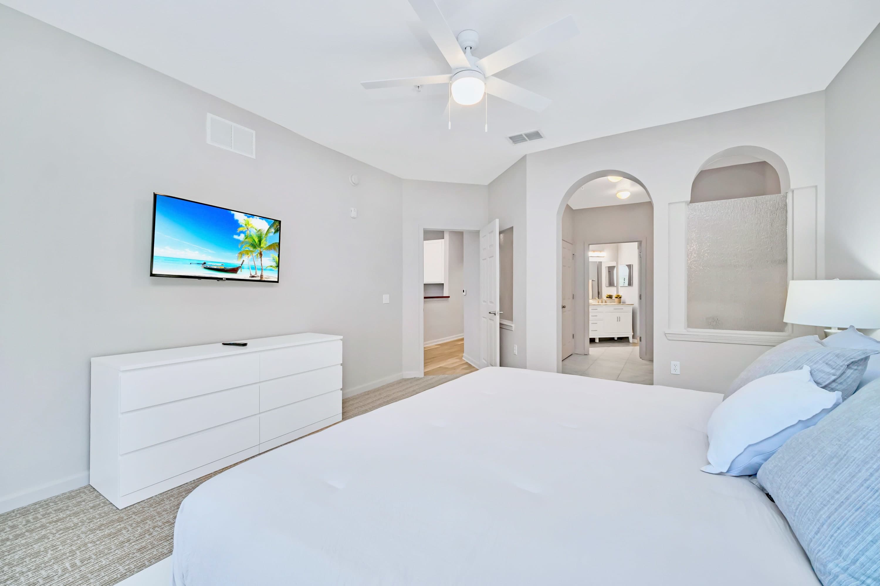 Spacious master bedroom with a white bed, matching dresser, and a wall-mounted TV, featuring light gray walls and an arched entryway leading to the bathroom.