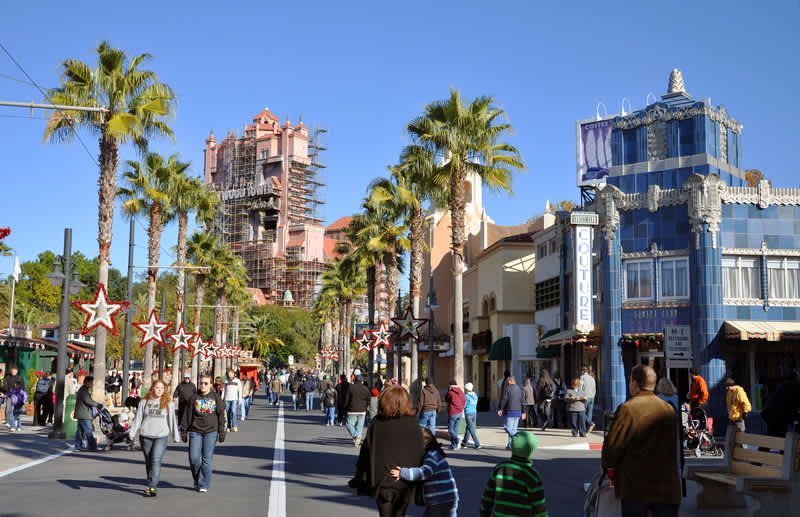 Bustling street scene at a theme park, resembling Hollywood Studios, with people walking along a wide boulevard lined with palm trees and themed buildings. The Tower of Terror ride is visible in the distance.