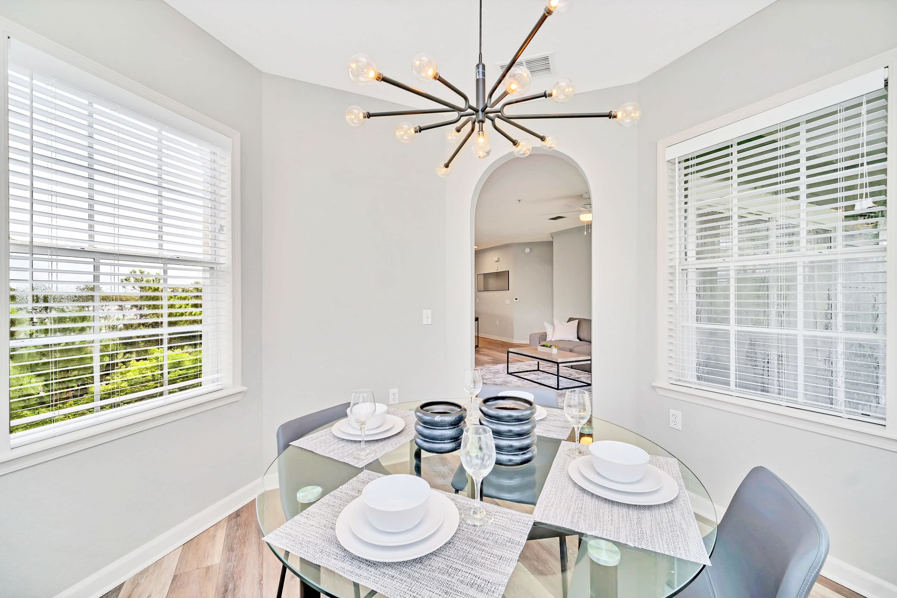 Close-up of the dining nook with a round glass-top table set for four, featuring white dinnerware and modern decor. The sputnik-style chandelier hangs above, and multiple windows with white blinds surround the space.