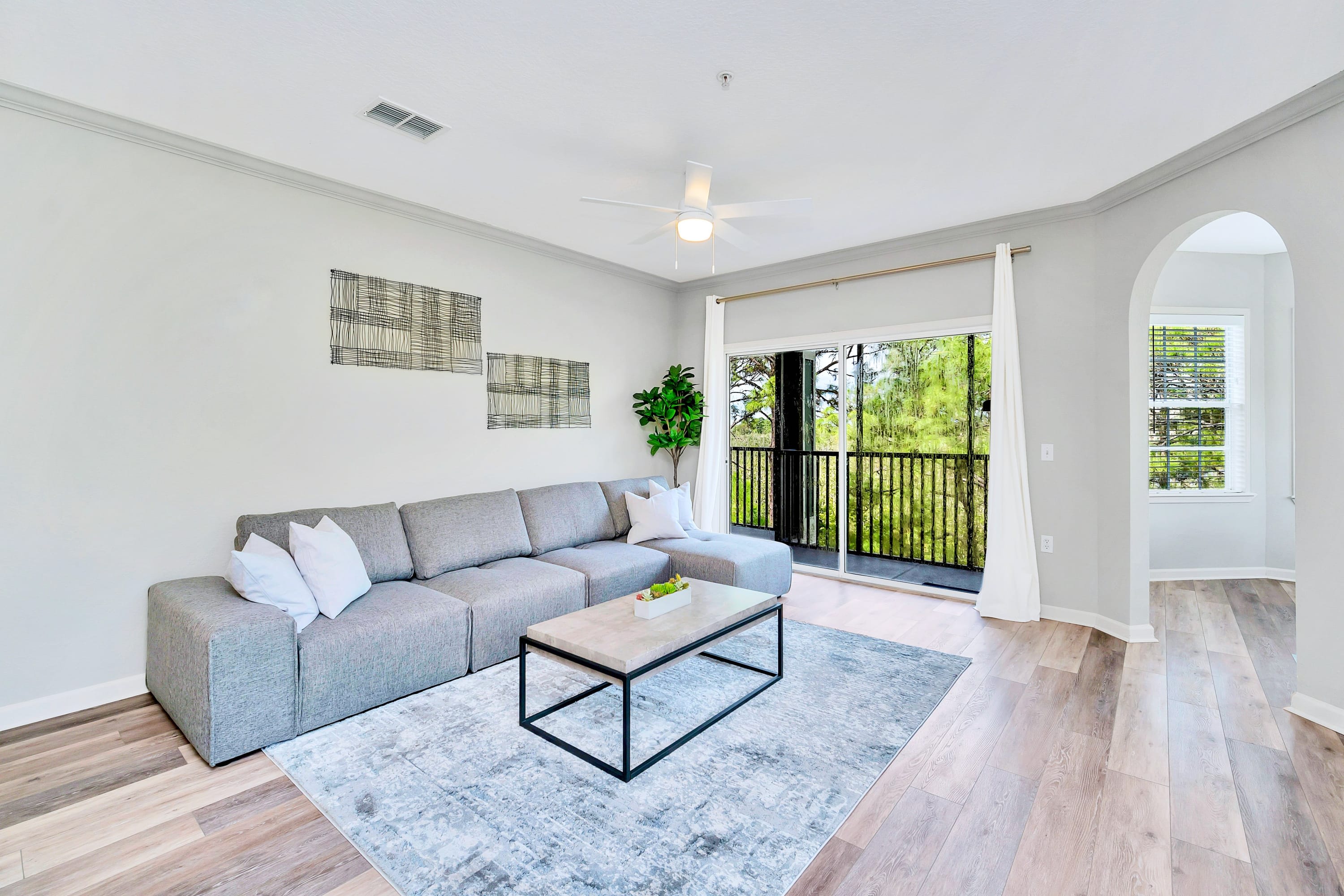 Wide view of a modern living room featuring a large L-shaped gray sectional sofa with white throw pillows. A rectangular coffee table sits on a patterned rug, and a potted plant adds greenery, with a sliding glass door leading to a balcony.