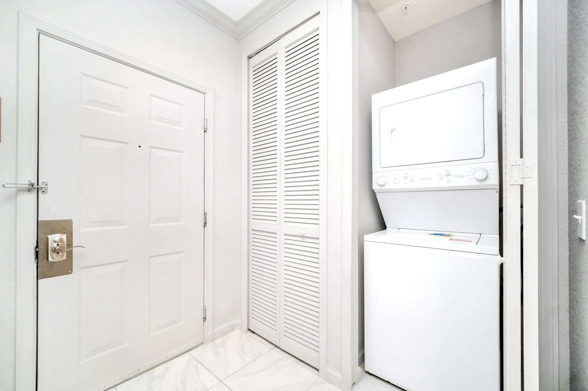 Compact laundry area with a stacked white washer and dryer unit, located near the entrance door and a louvered closet, on a white tiled floor.