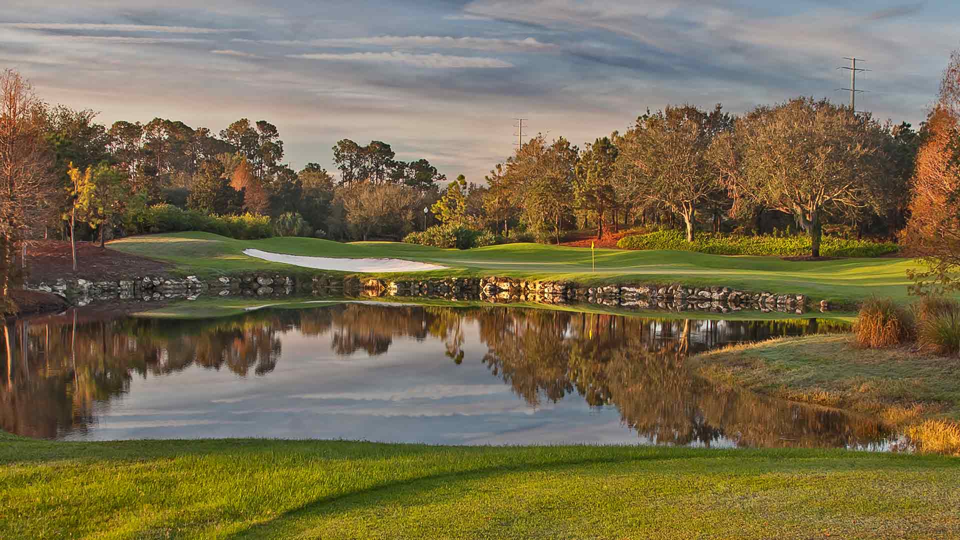 Scenic view of a golf course hole featuring a green flag, a sand bunker, and a calm pond reflecting the cloudy sky and surrounding trees. The foreground shows manicured green grass.
