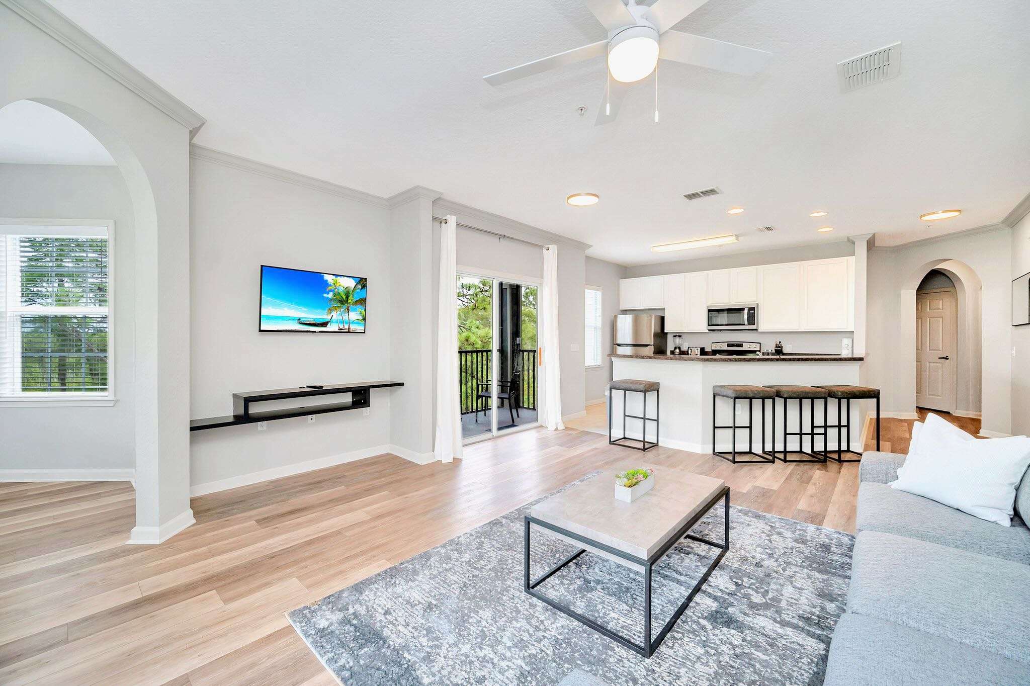 Bright living room with light wood-look flooring, a wall-mounted TV, and a sliding glass door leading to a balcony. A breakfast bar with stools separates the living area from the kitchen, which has white cabinets and stainless steel appliances.