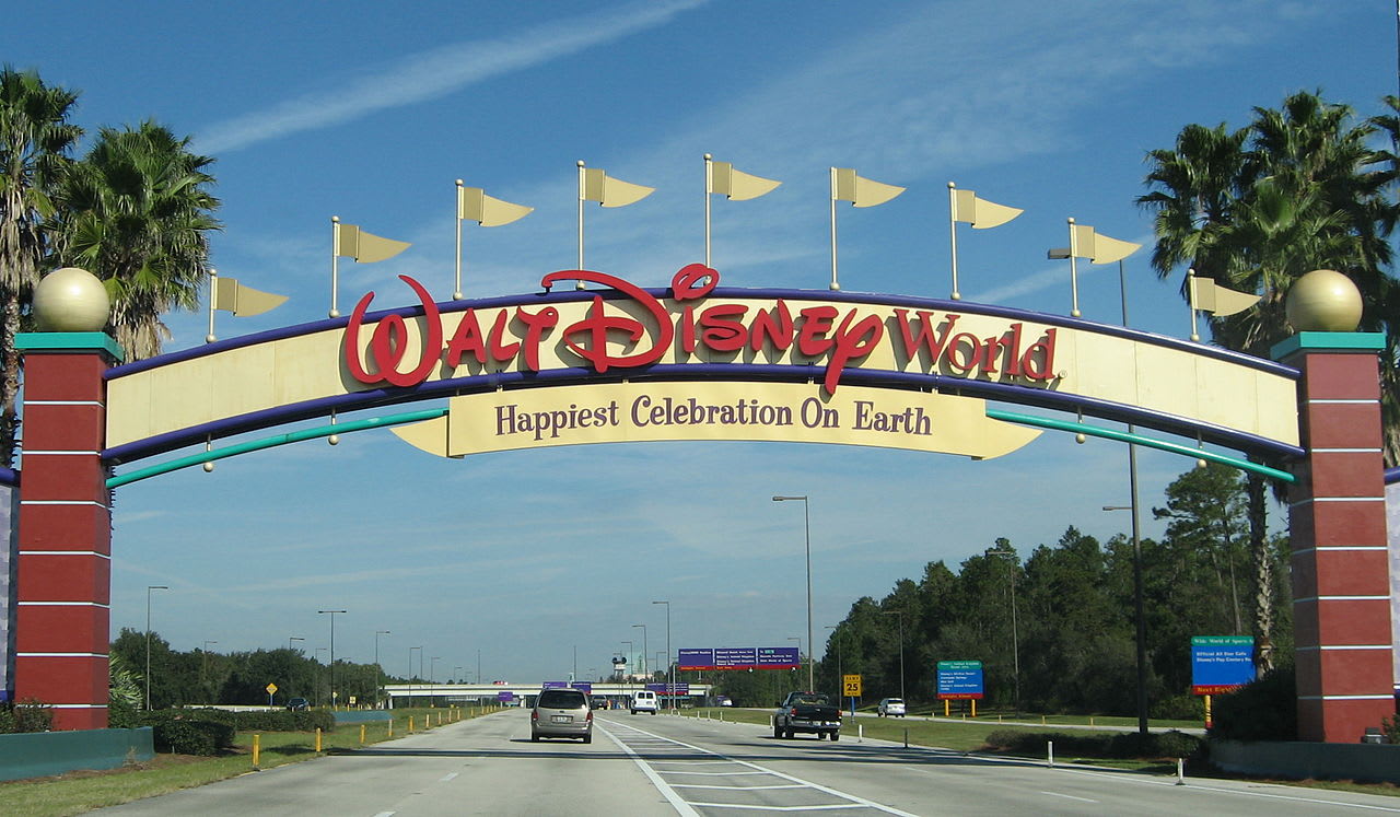 Iconic Walt Disney World entrance archway spanning a multi-lane highway, featuring the "Happiest Celebration On Earth" slogan and golden flags. Palm trees line the road under a bright blue sky.