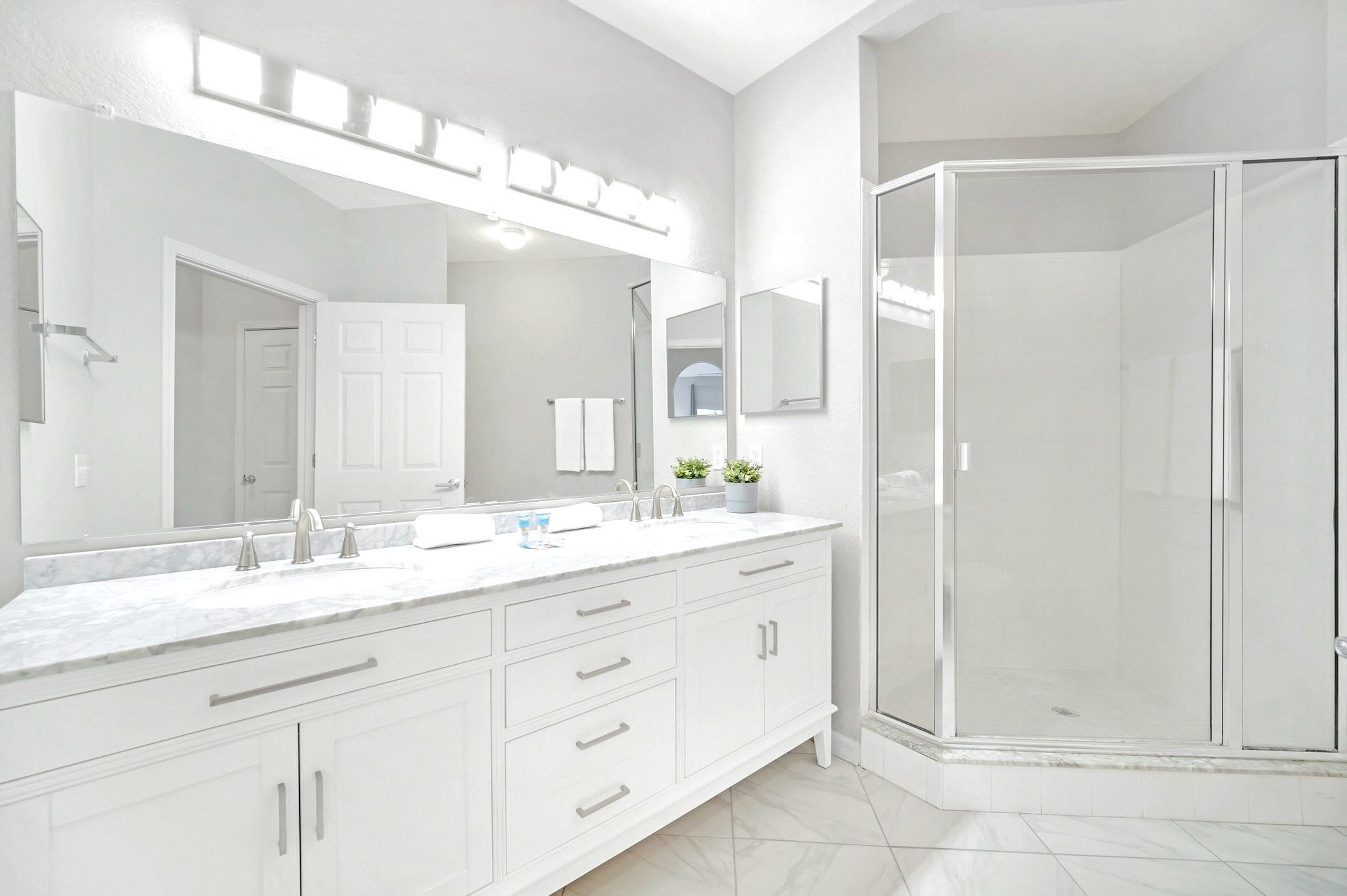 Modern master bathroom featuring a long white double vanity with marble-look countertops, a large mirror, and a glass-enclosed corner shower, all on a white tiled floor.