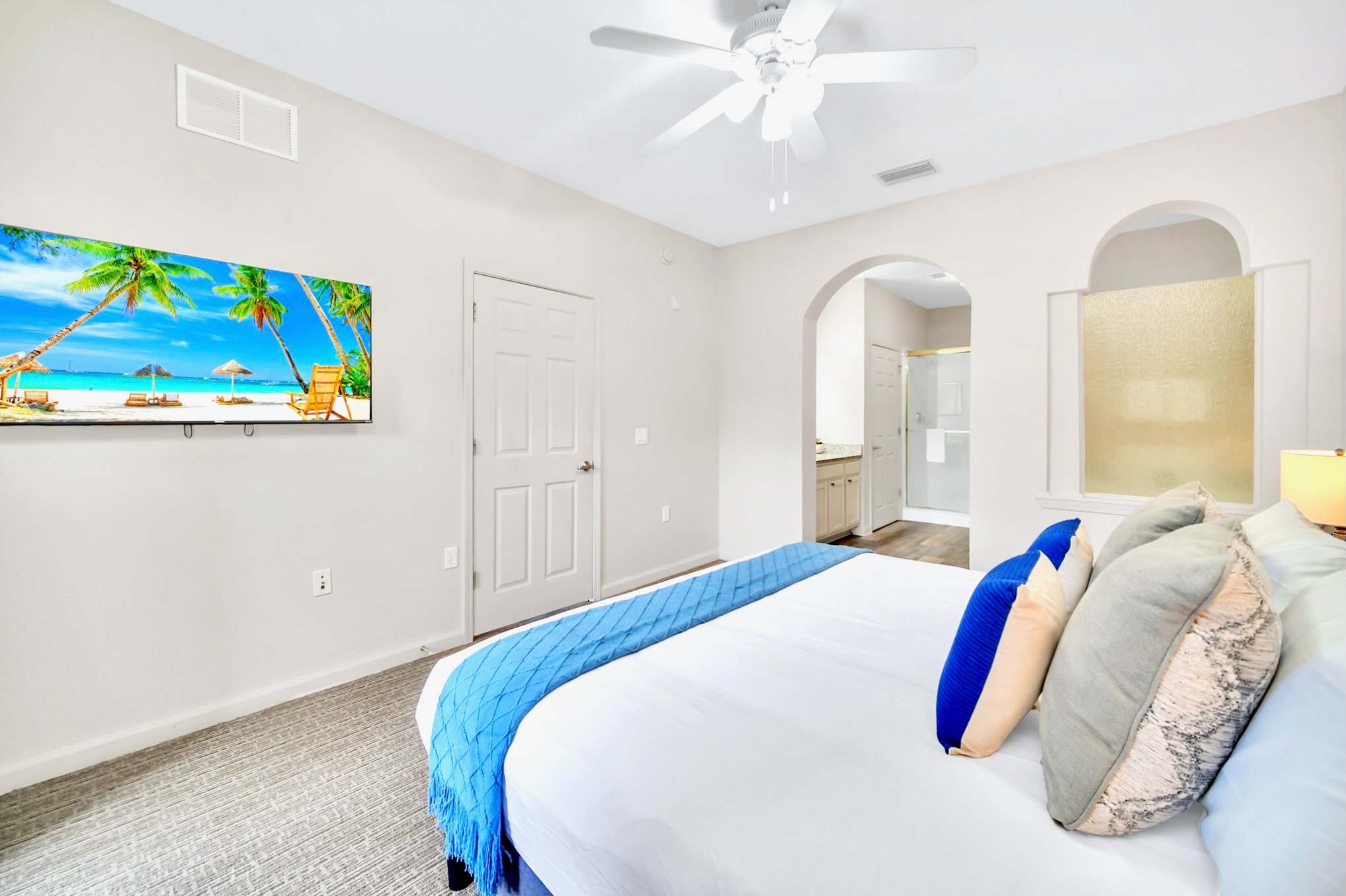 Master bedroom with a king-sized bed dressed in white and blue bedding, light gray carpet, and a wall-mounted TV displaying a beach scene. An arched doorway leads to the en-suite bathroom, revealing a vanity area.