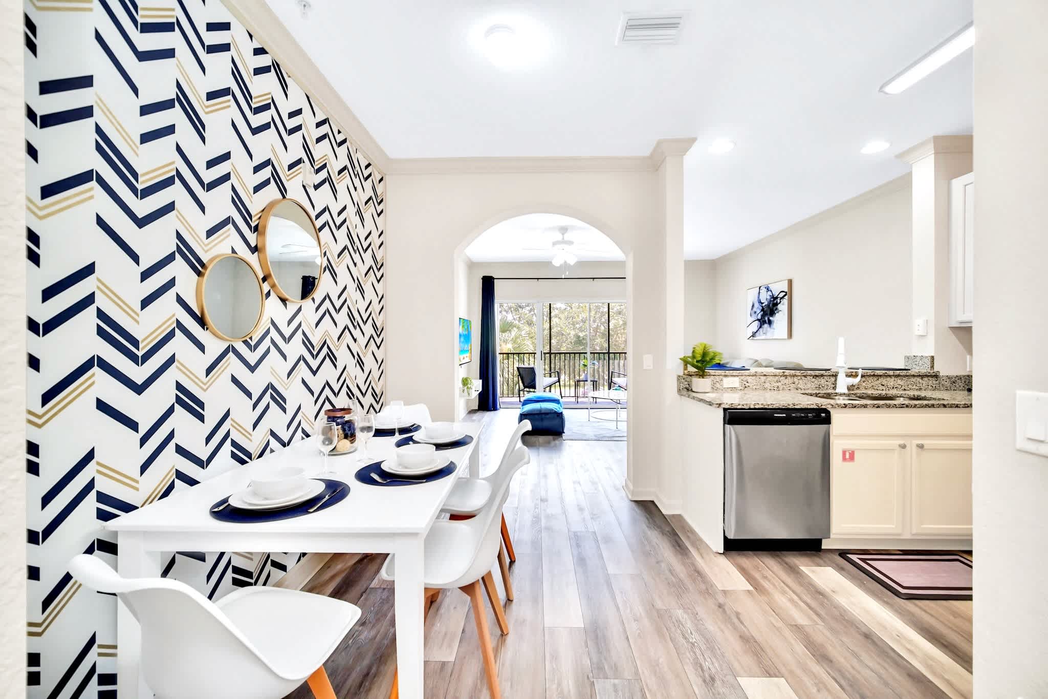 Open-concept view showing the dining area with its chevron accent wall, leading into a modern kitchen with light cabinetry, granite countertops, and stainless steel appliances. An arched entryway connects to the living space.
