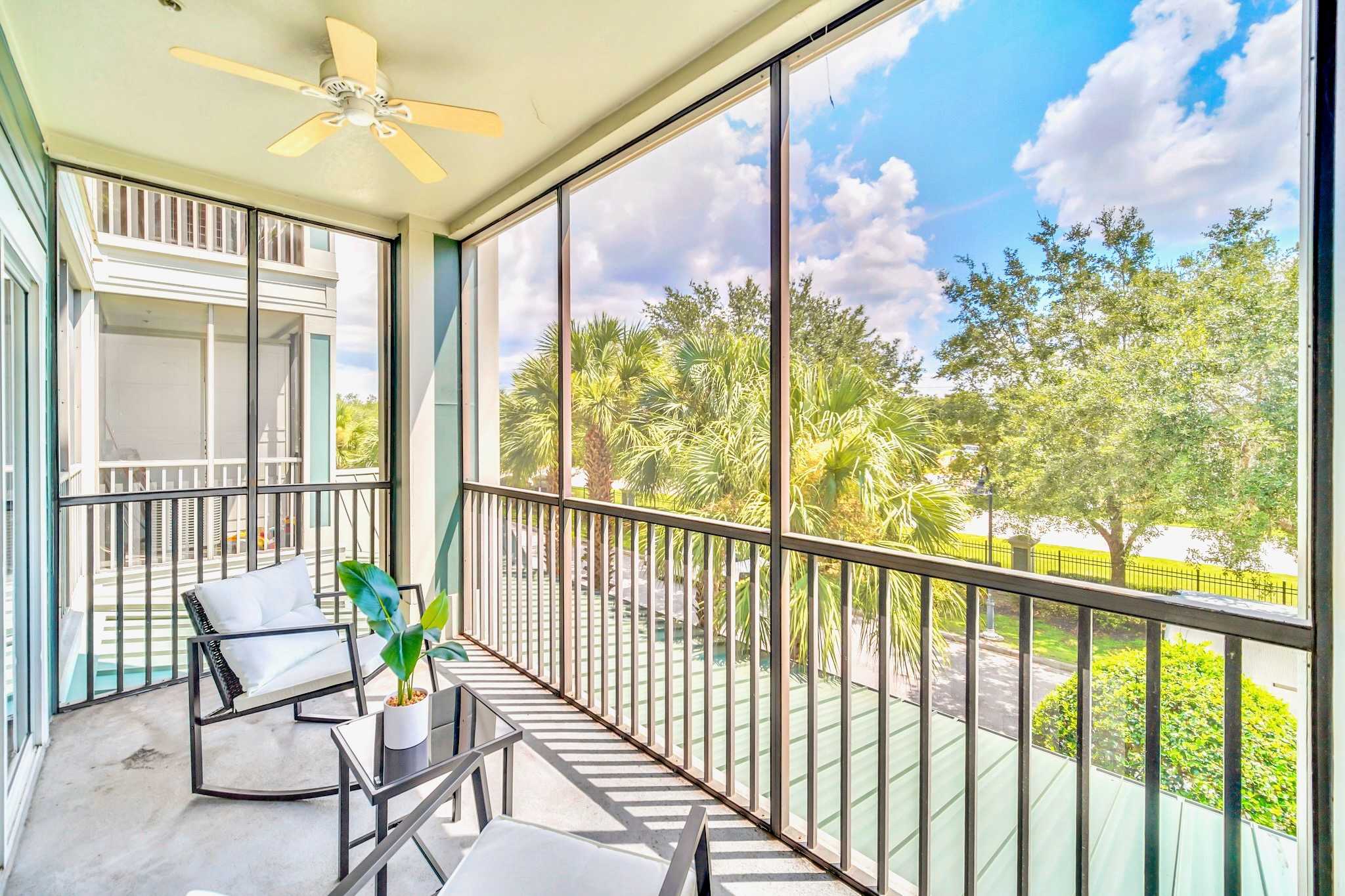 Screened balcony with a light-colored ceiling fan, furnished with two black metal chairs and a small side table. The balcony offers expansive views of lush trees and a body of water under a bright blue sky with clouds.