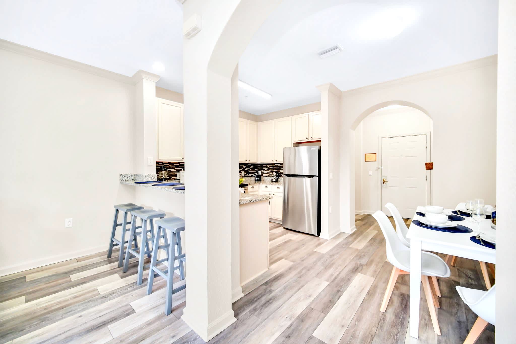 View of the kitchen bar with three gray bar stools and the adjacent dining area featuring a white table and chairs, all set on light wood-look flooring. The kitchen is visible in the background through an arched opening.