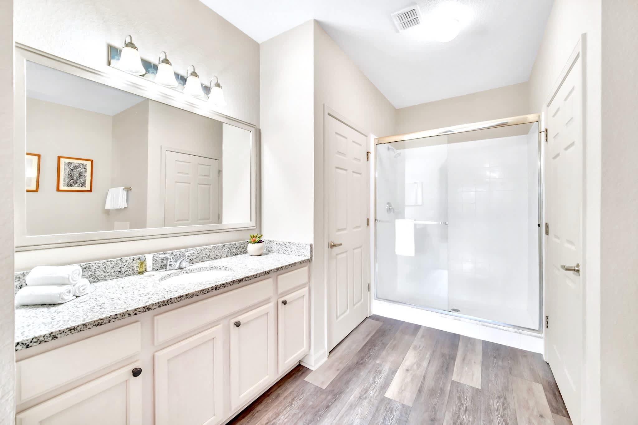 En-suite bathroom with a large vanity featuring a granite countertop, single sink, and a wide mirror with decorative lighting. A glass-enclosed walk-in shower is visible to the right, and light wood-look flooring covers the floor.