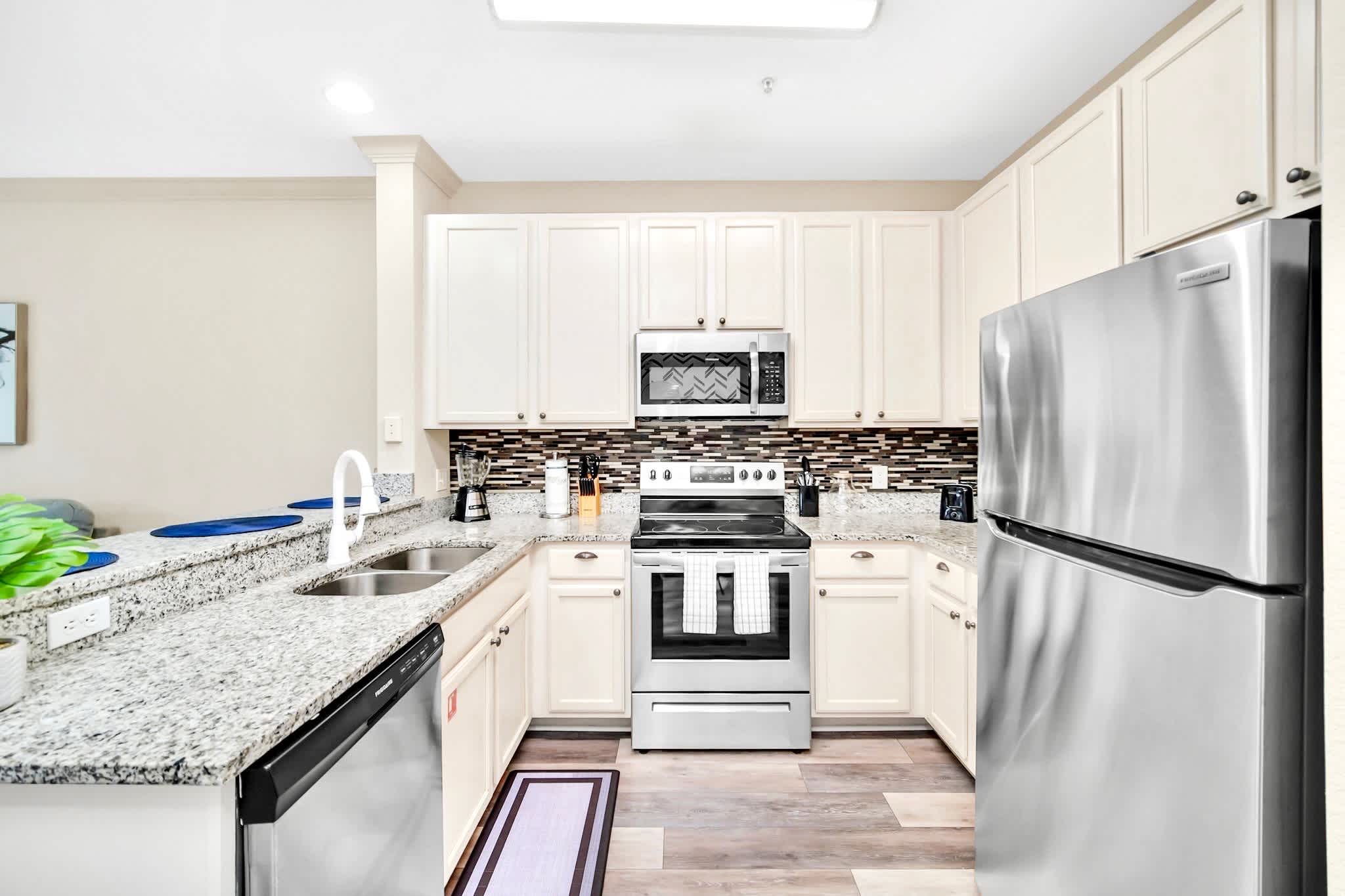 Full view of the modern kitchen featuring light cream cabinetry, granite countertops, and stainless steel appliances including a refrigerator, range, microwave, and dishwasher. A mosaic tile backsplash adds texture, and light wood-look flooring extends throughout.
