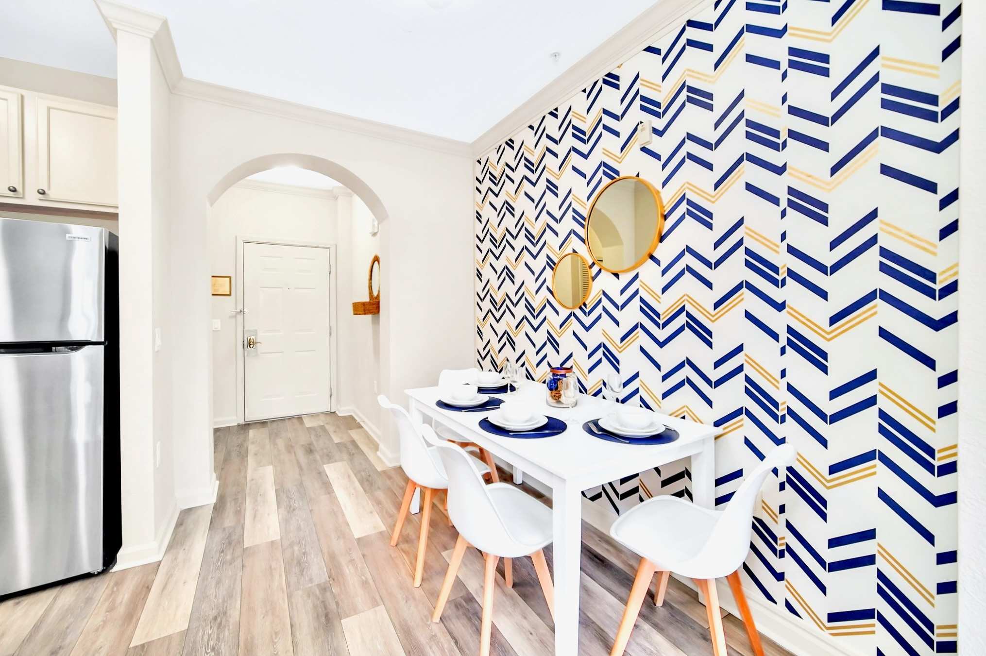 Dining area with a white table and chairs, featuring a bold chevron accent wall with blue, gold, and white patterns. Beyond the dining area, an arched doorway leads to the front entrance with a white door and a woven mirror.