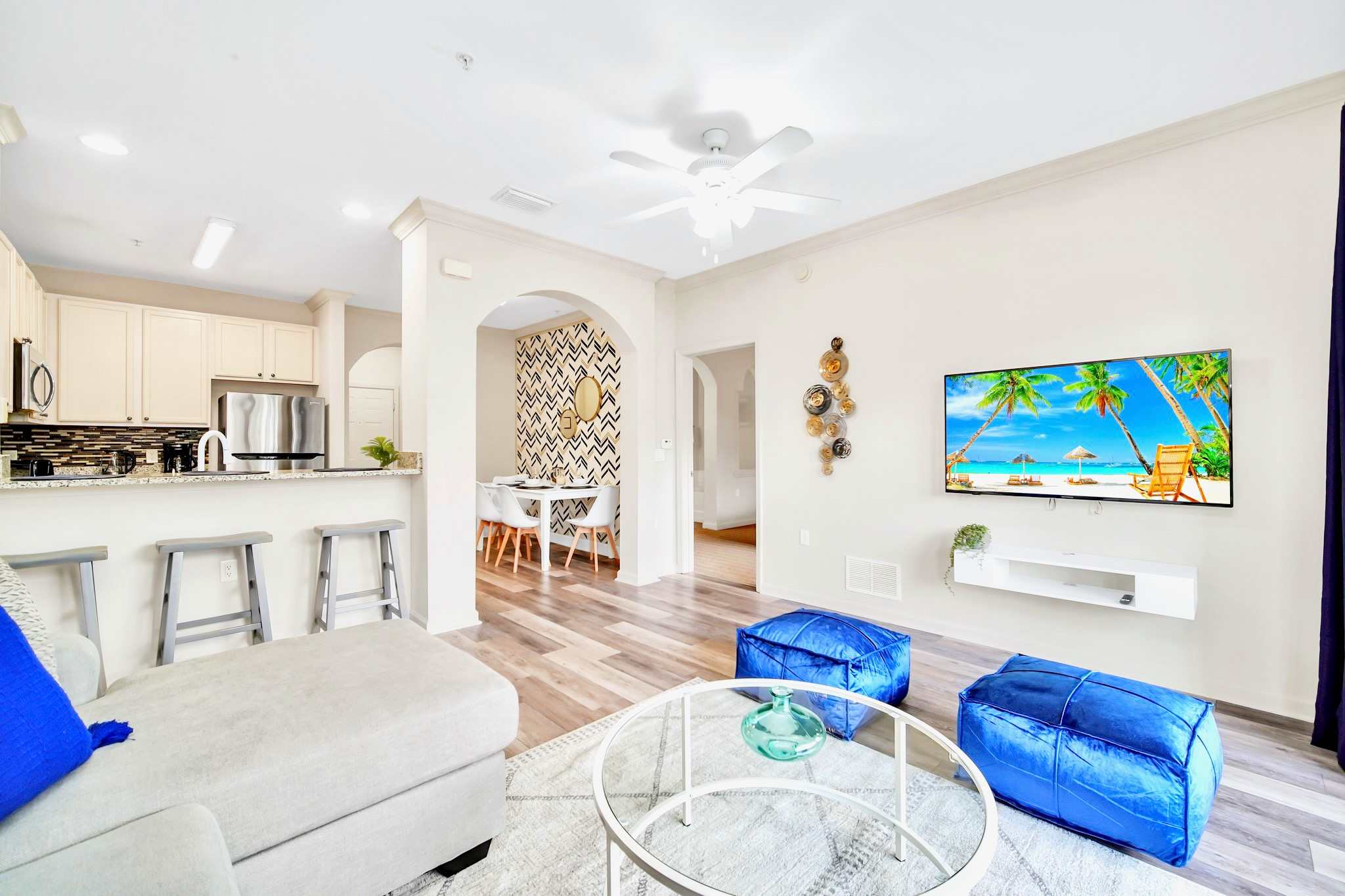 Bright open-concept living space with light wood-look flooring, a light gray sectional sofa, a glass coffee table, and two blue poufs, with a wall-mounted TV displaying a beach scene. The kitchen bar and dining area are visible through arched doorways.
