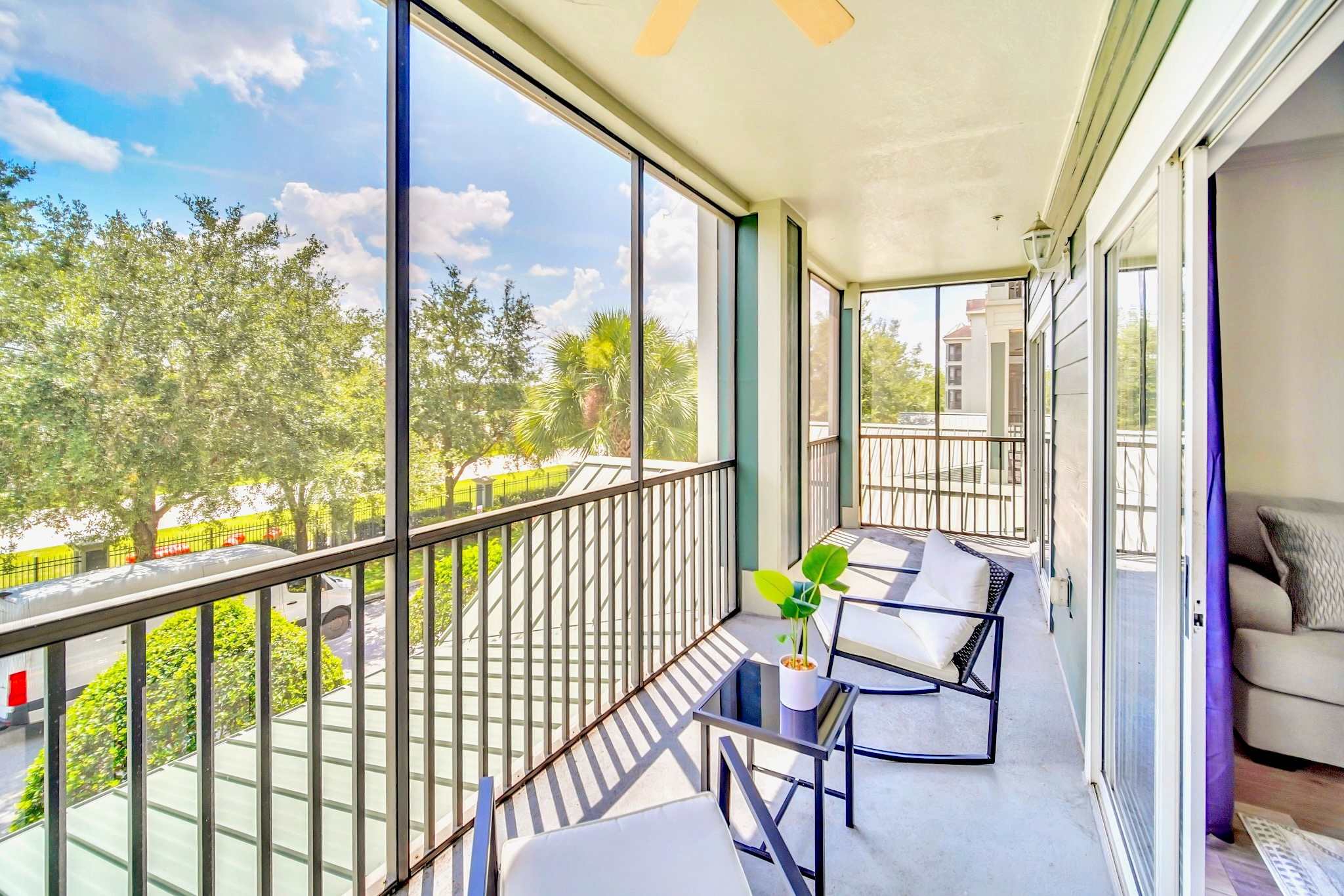 Wide view of a screened balcony, showing two black metal chairs and a side table, with a sliding glass door leading into the interior. The balcony overlooks a scenic landscape of trees and a body of water under a bright sky.