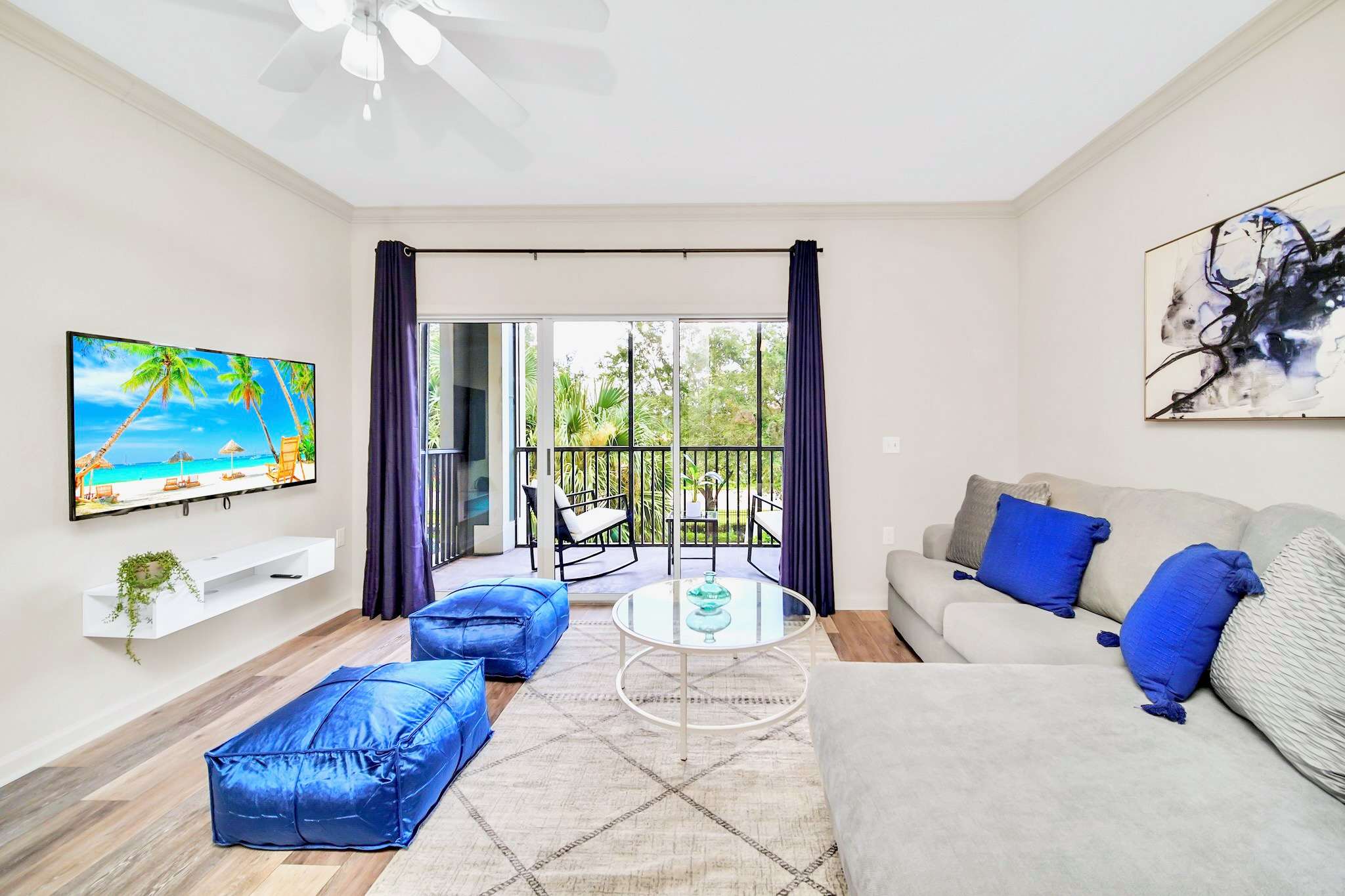 Spacious living room with light wood-look flooring, a light gray sectional sofa with blue accent pillows, a glass coffee table, and two blue poufs, facing a wall-mounted TV. A sliding glass door with dark blue curtains opens to a balcony with outdoor seating.