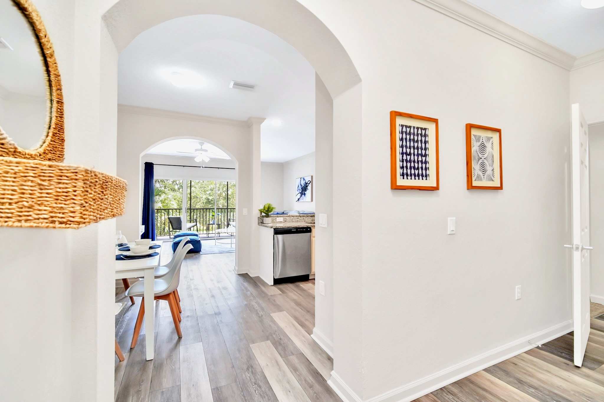 Hallway with light hardwood floors and white walls, featuring two framed artworks. An arched opening leads to the open-concept living area and kitchen, with a glimpse of the dining area to the left.