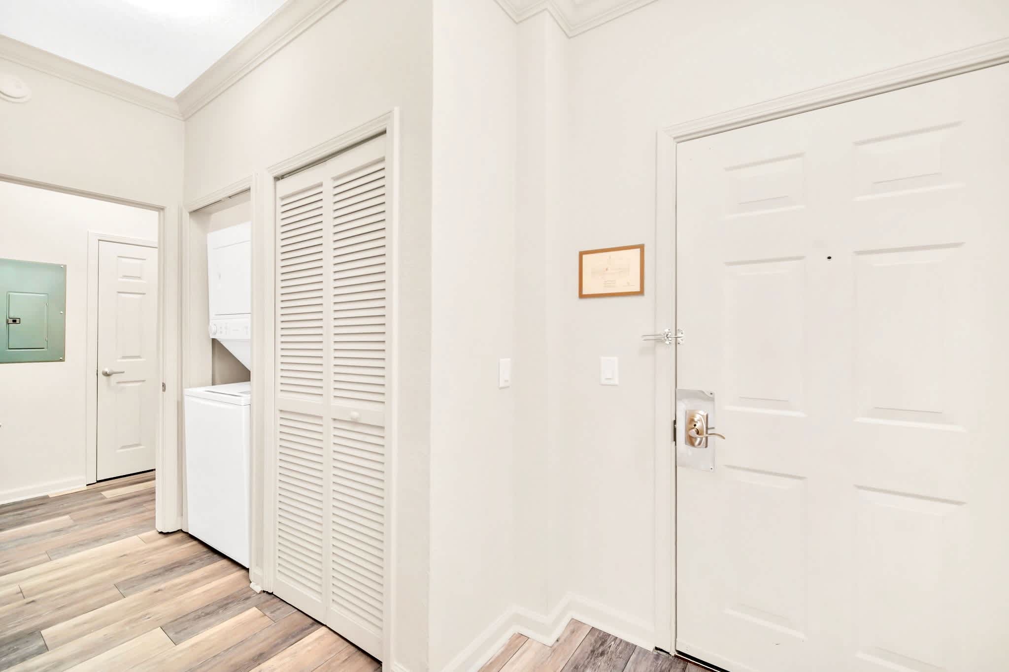 A bright and clean foyer with light-colored walls and wood-look laminate flooring, featuring a louvered bifold door revealing a stacked washer and dryer unit. A white entry door with a silver handle is on the right.