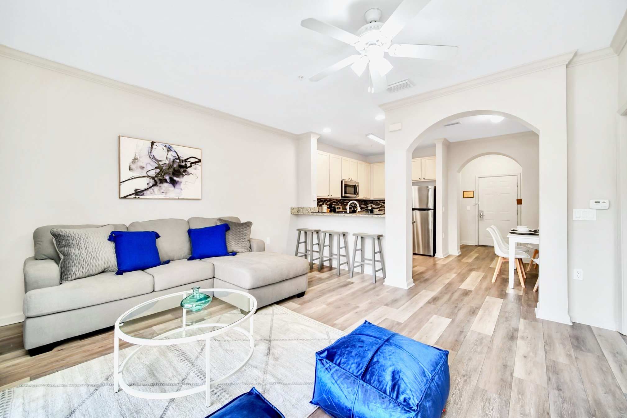 Open-concept living room with a light gray sectional sofa and blue pillows, facing a kitchen bar with three gray bar stools. The kitchen features light cabinetry and stainless steel appliances, visible through an arched opening.